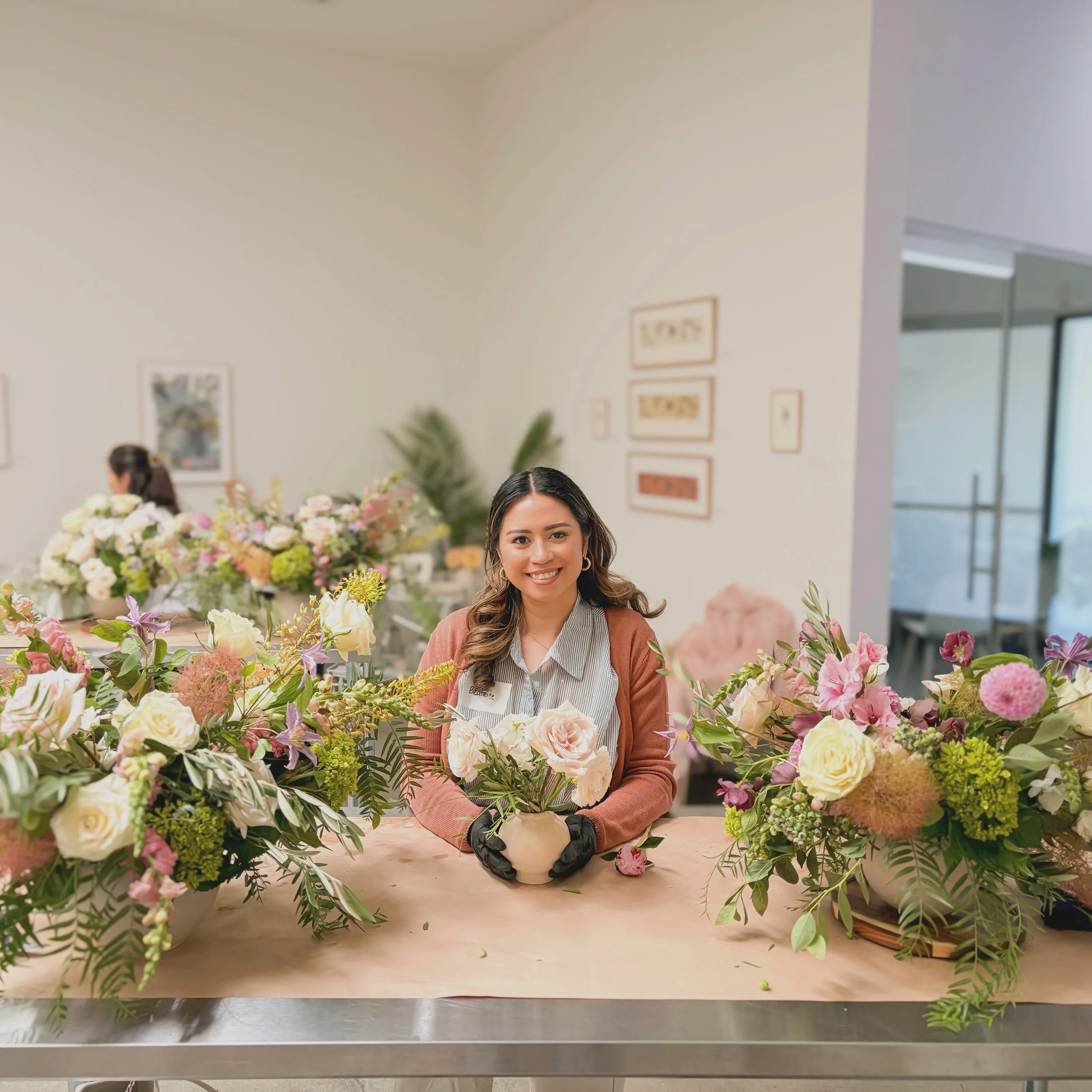 A woman with long brown hair, wearing a brown jacket and black gloves, is sitting at a table filled with a large variety of pink, white, cream, and purple flowers and greenery. There are framed pictures hanging on the white wall behind her, and another woman can be seen in the background.