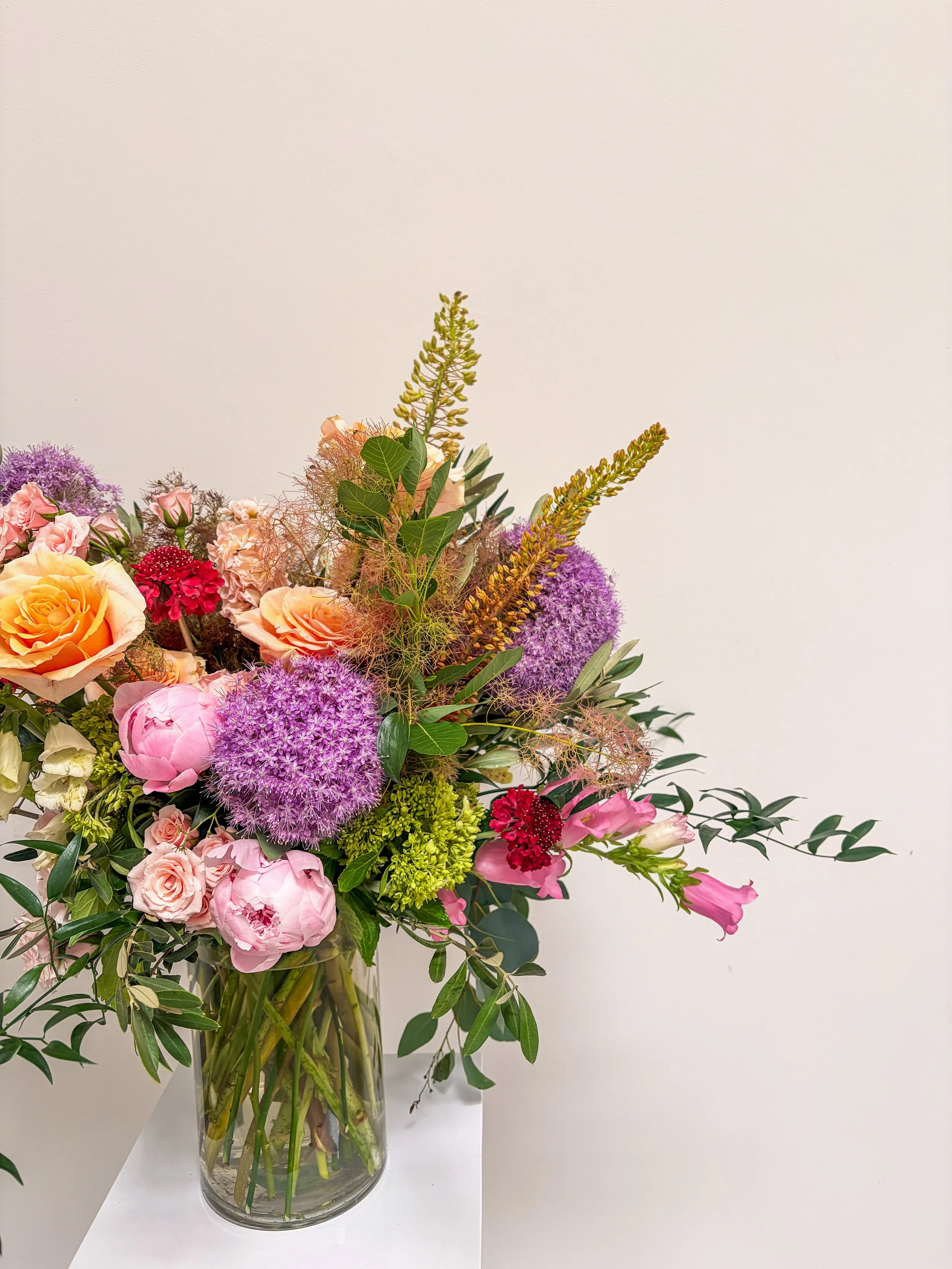 A large bouquet of mixed colorful flowers in a tall, clear glass vase on a white surface against a plain off-white wall.