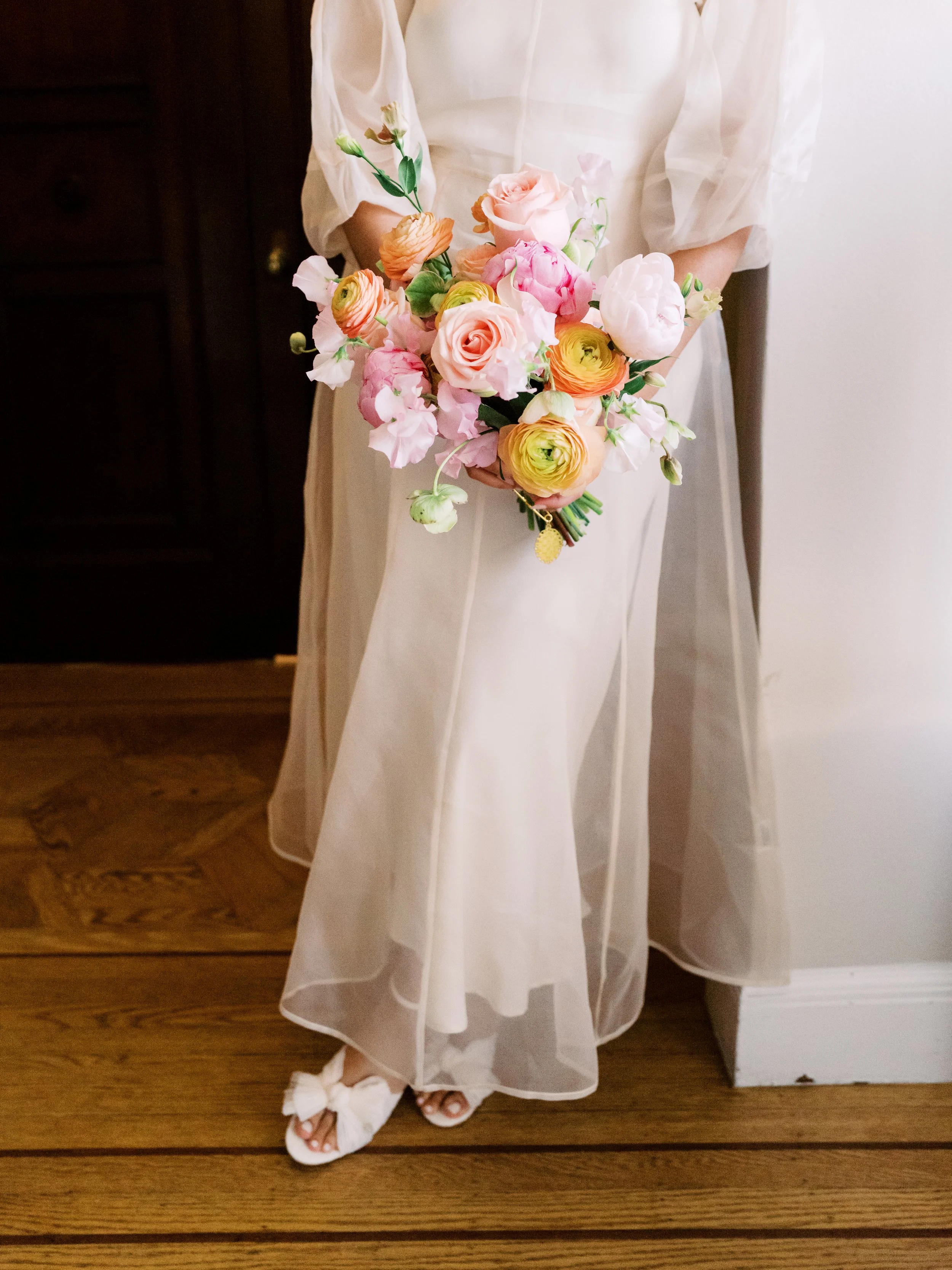 A woman in a long, flowing white dress holding a colorful bouquet of pink, yellow, and white flowers. The dress is sheer, with puffed sleeves, and she is wearing white floral sandals.