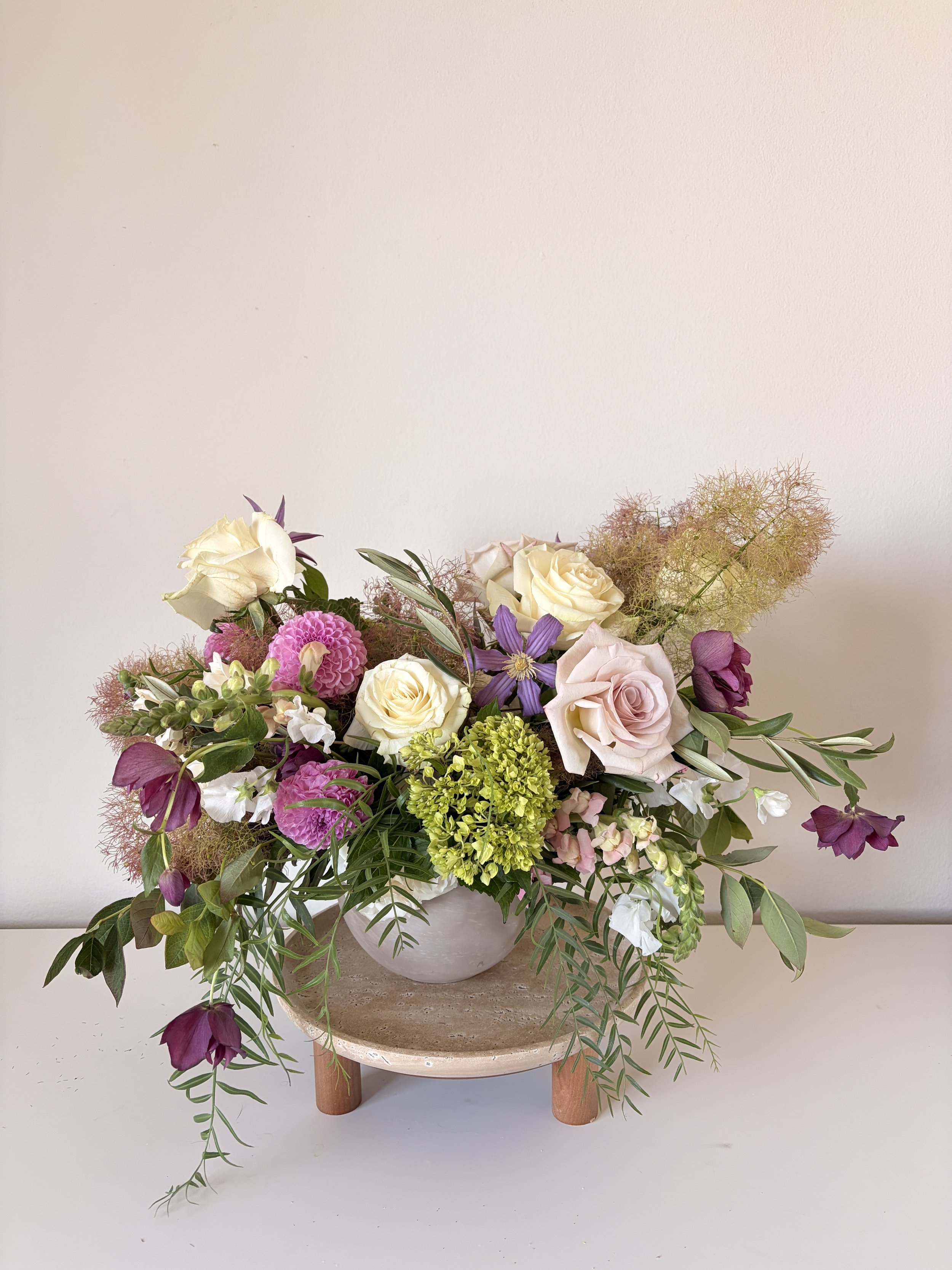 A floral arrangement in a white vase with pink, white, purple, and green flowers and greenery, placed on a round wooden stand against a plain white wall.
