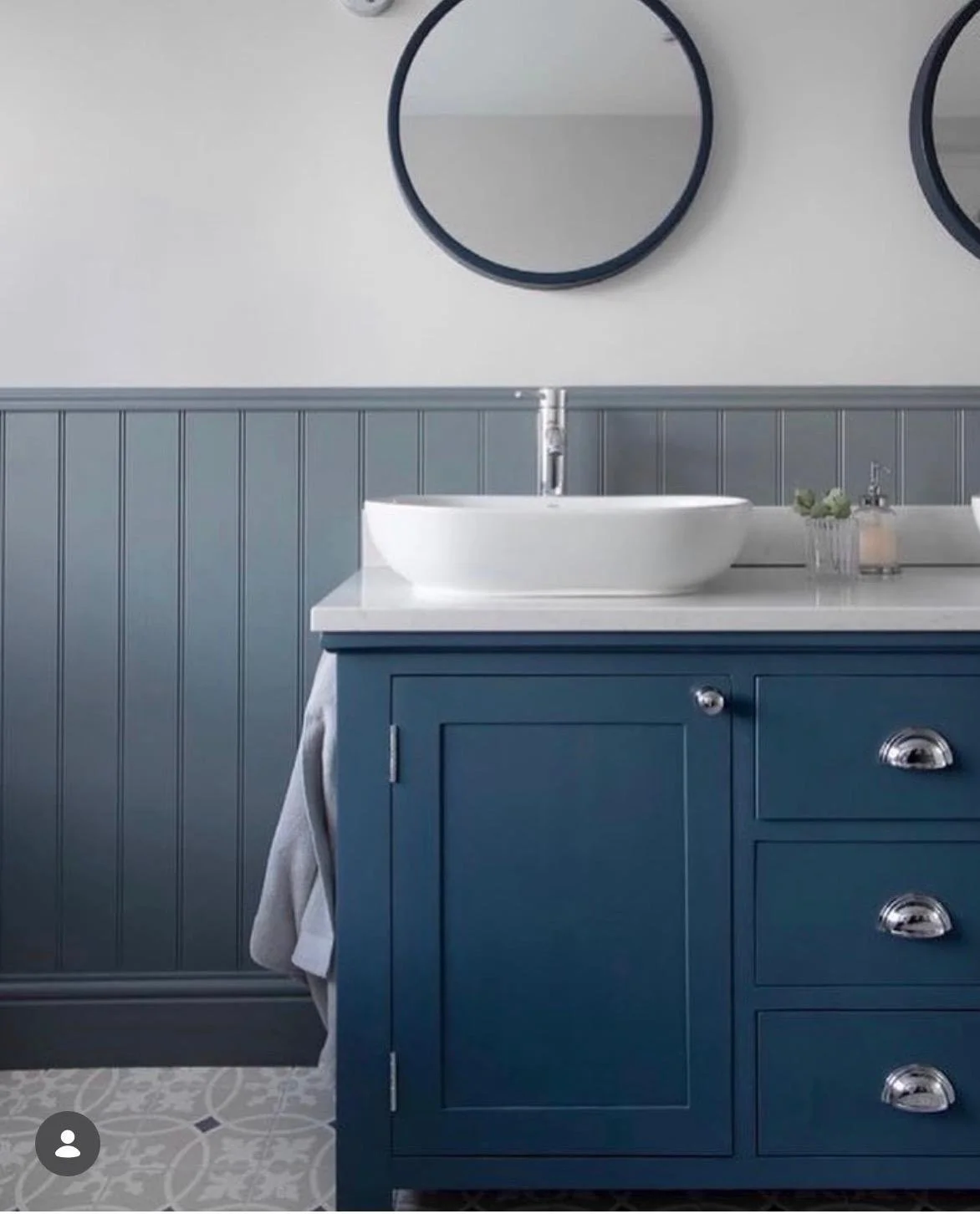 A bathroom vanity with a white vessel sink, blue cabinets, a gray beadboard wall, and a round mirror above.