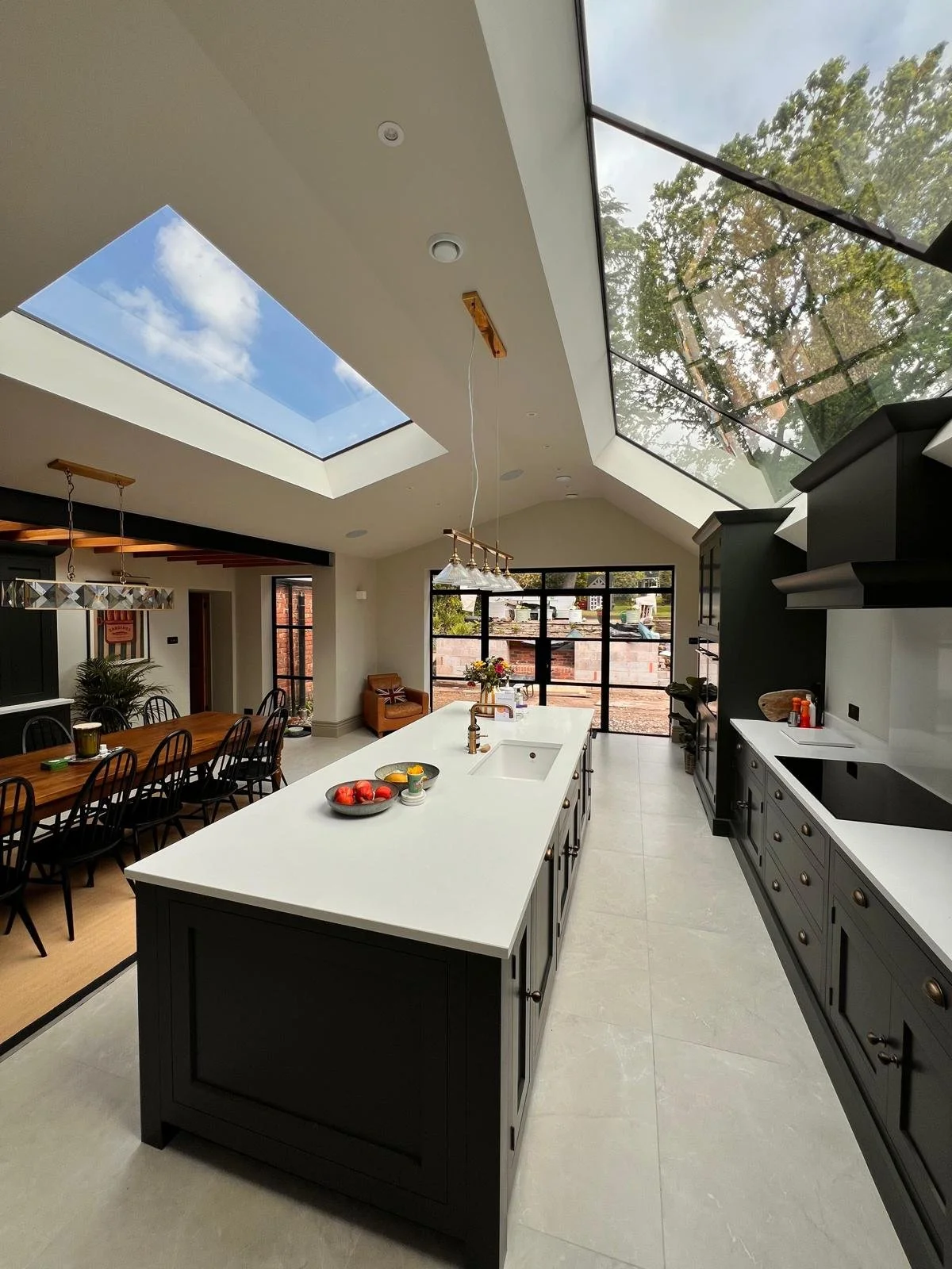 Modern kitchen with a large white island, black cabinetry, and a skylight showing a blue sky with clouds. There is a dining area with a wooden table and black chairs, and large windows overlooking a construction site outside.