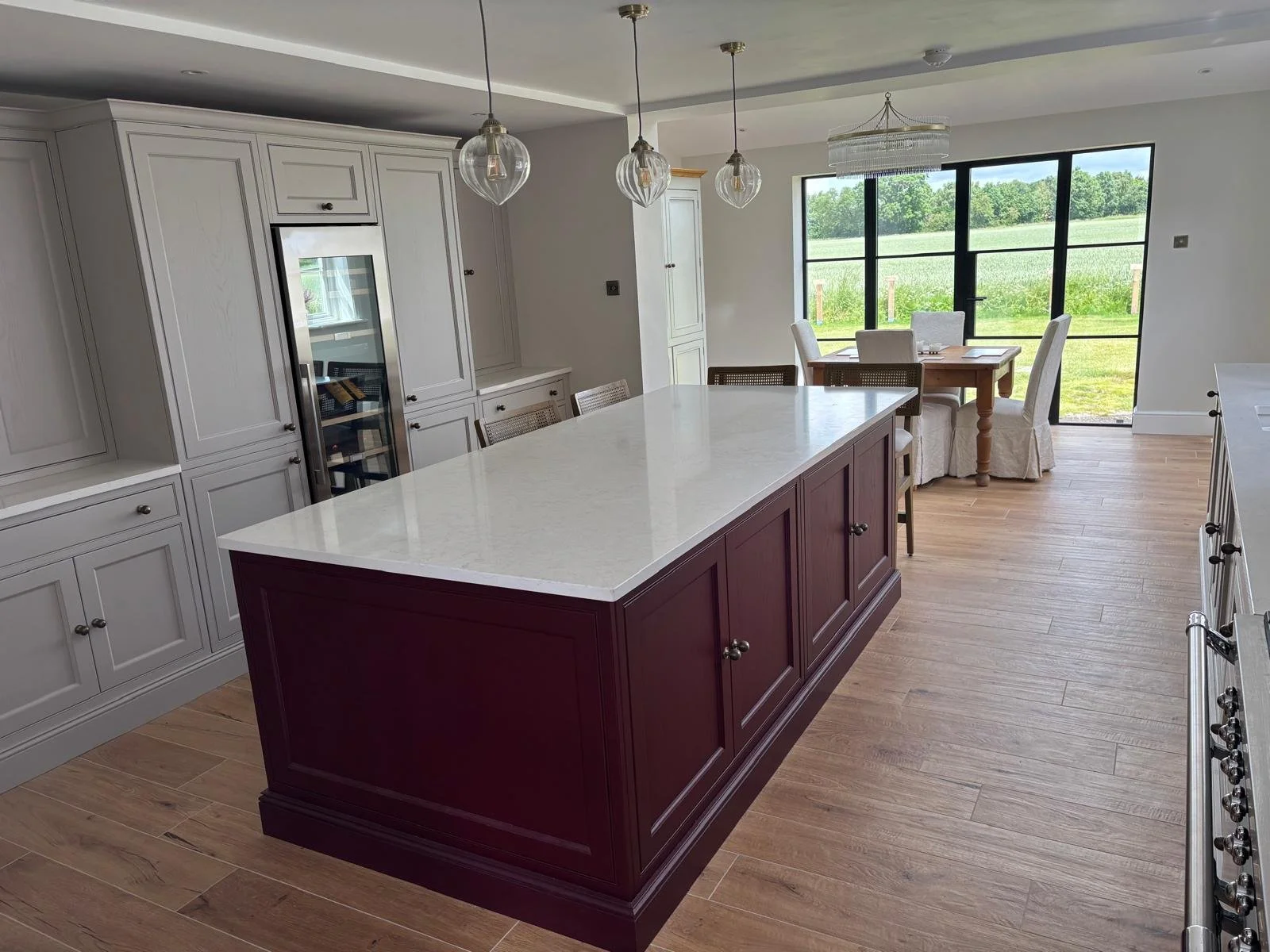 Modern kitchen with an island that has a maroon base and white countertop, white cabinetry, a wine fridge, pendant lighting, and a dining area with a table and chairs near large sliding glass doors overlooking a green outdoor area.