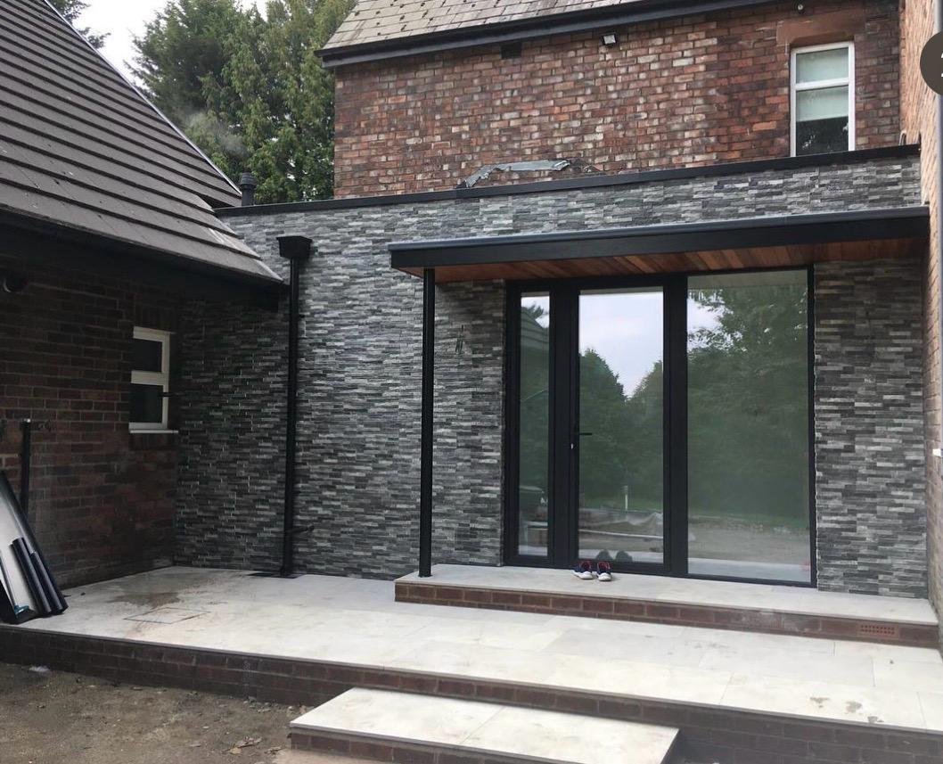 Newly constructed modern patio with large glass sliding door, gray stacked stone exterior wall, brick house with brick steps leading to patio, and sneakers on the ground.