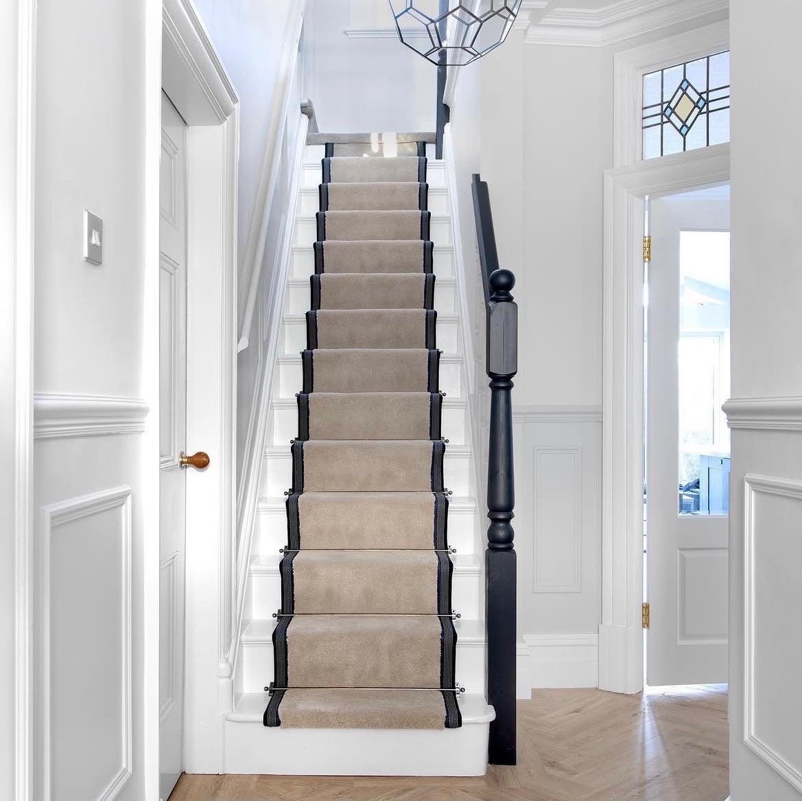 Interior view of a staircase with a beige runner and black binding, white walls, and a black handrail, in a bright, modern home.