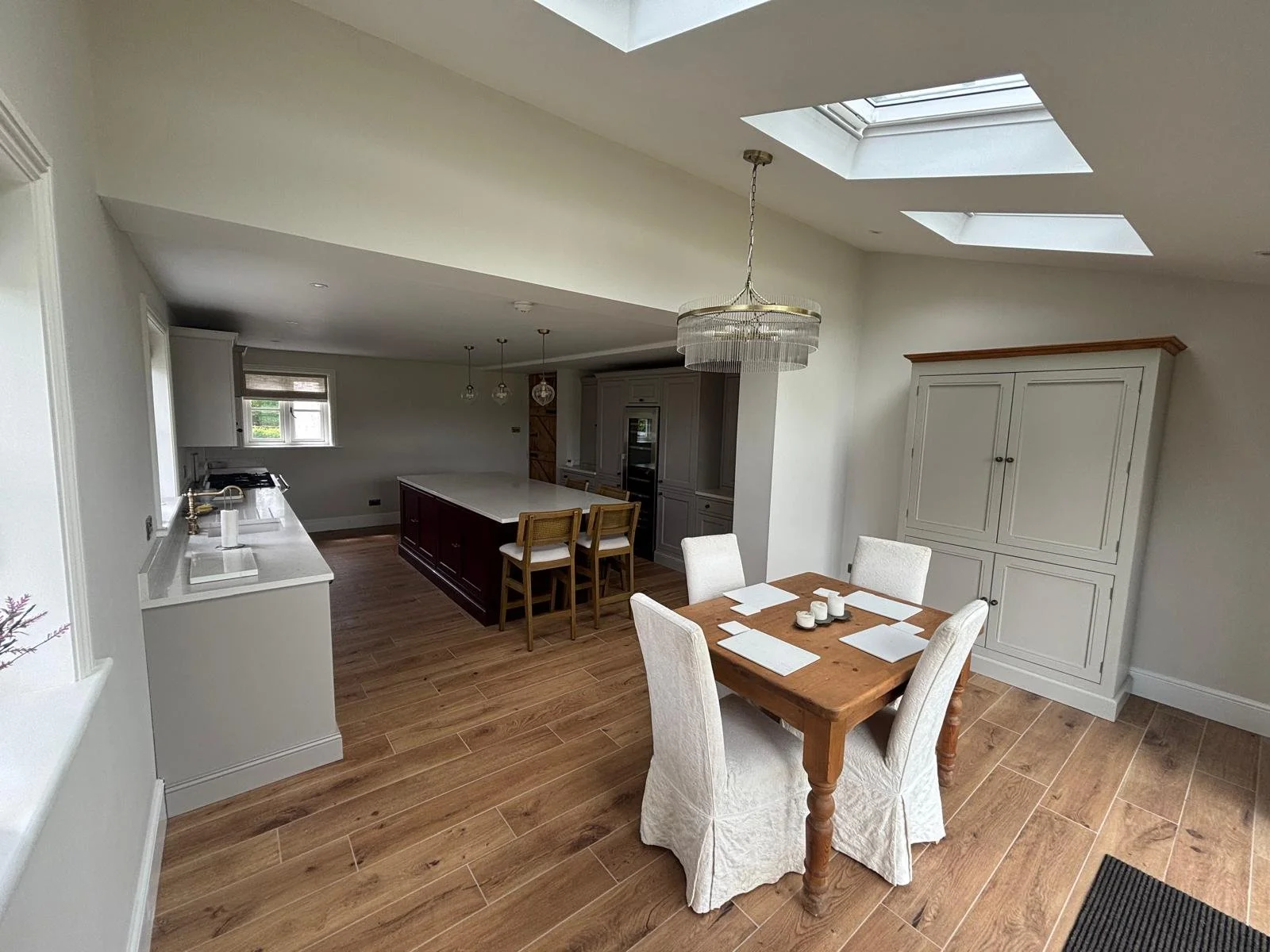 A spacious kitchen and dining area with natural light from skylights. There is a wooden dining table with six white upholstered chairs, a large kitchen island with wooden chairs, and white cabinetry. The room has light-colored walls and hardwood floo