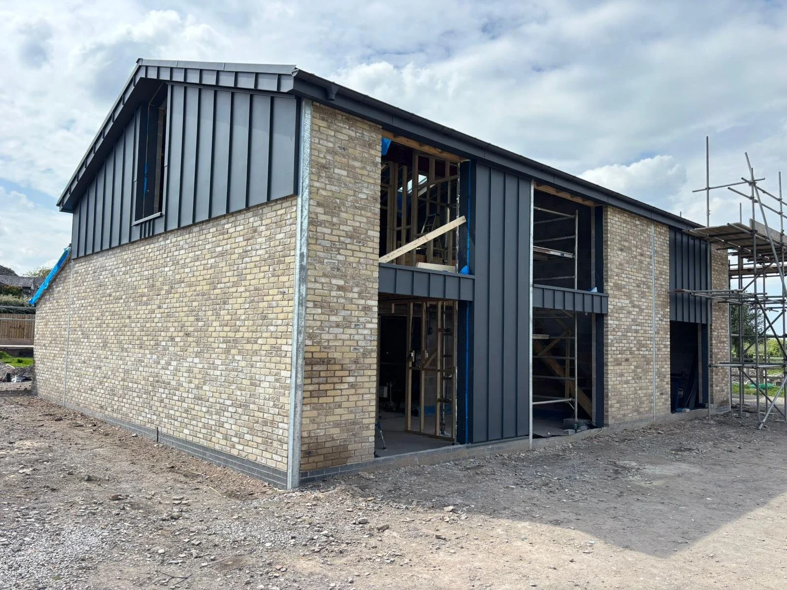 A two-story house under construction with brick walls, partially installed blue metal siding, and scaffolding on the right side, against a cloudy sky.