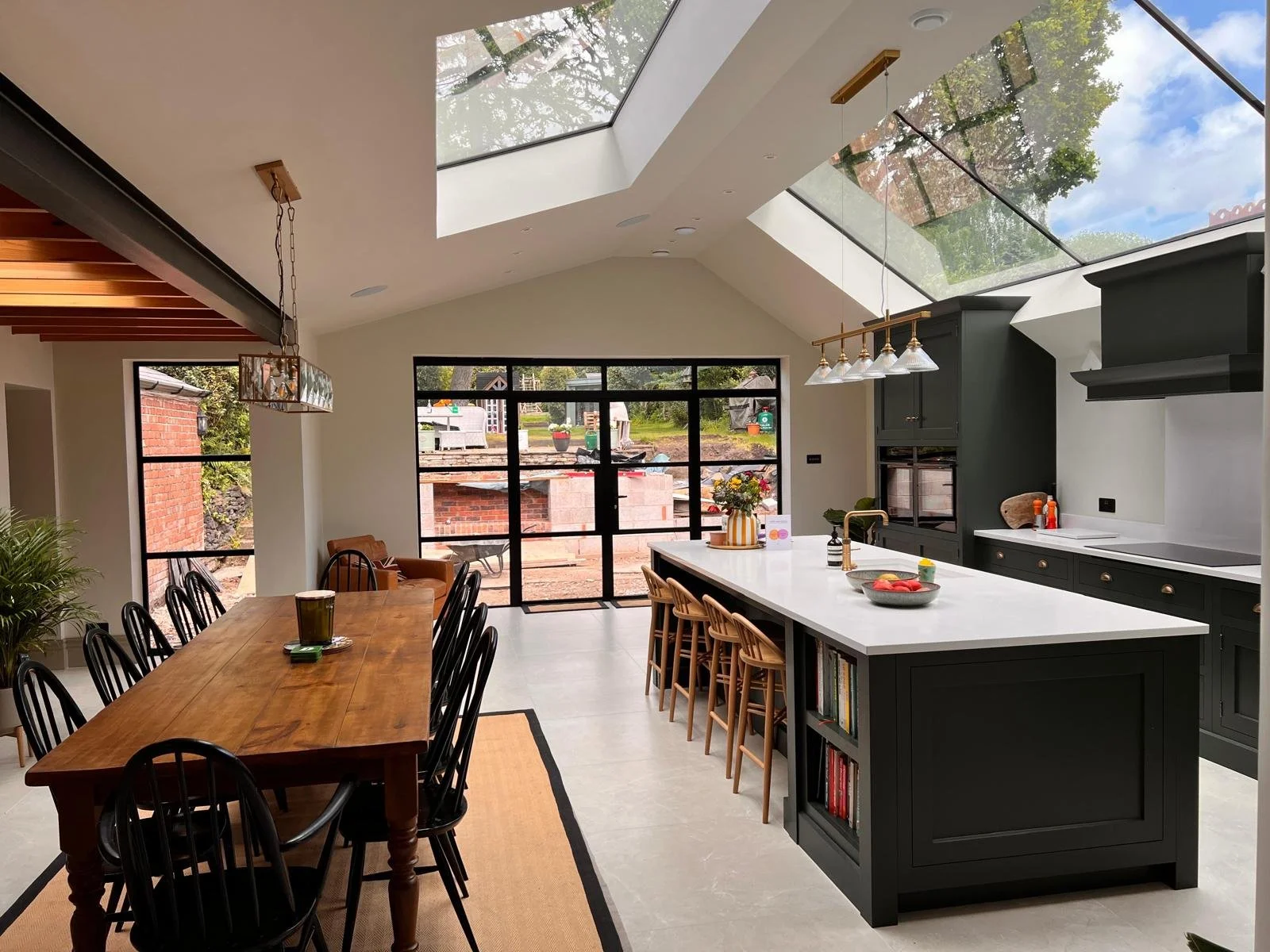 Modern kitchen and dining area with large skylight and glass doors, featuring a wooden dining table with black chairs, a black kitchen island with barstools, and dark cabinets, sunlight streaming in.
