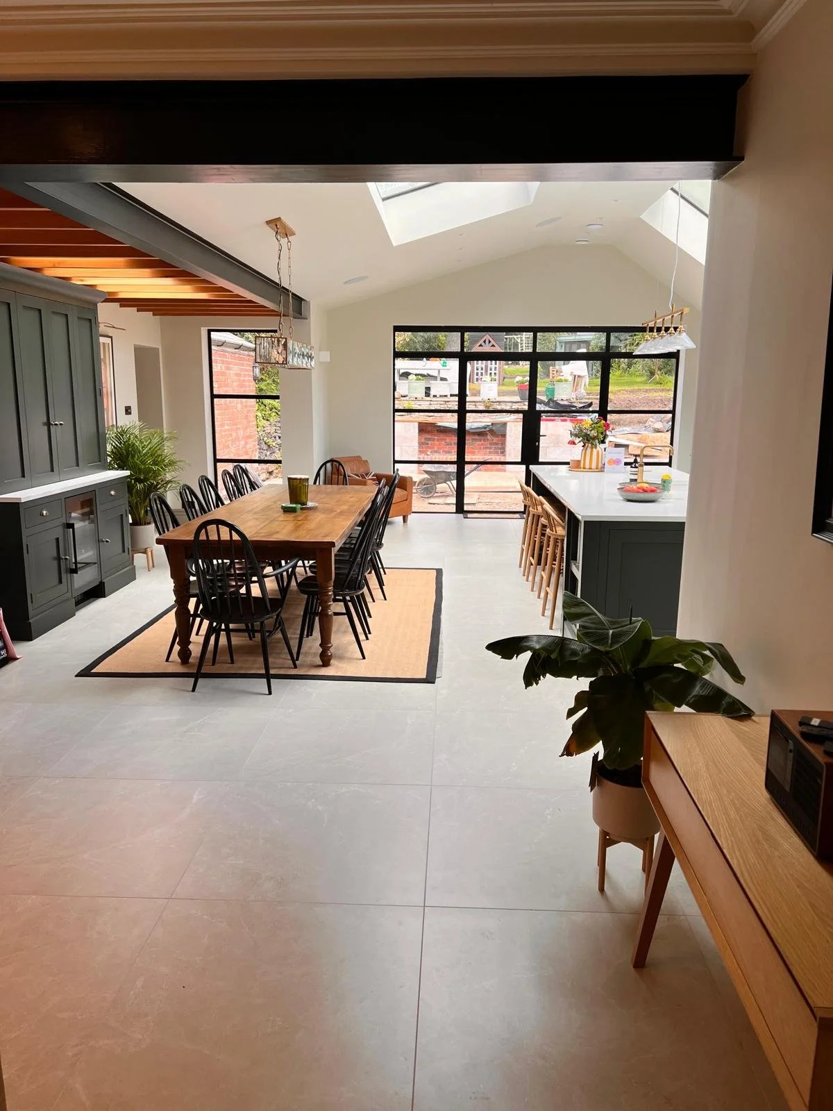 Open-concept dining area with a wooden table surrounded by black chairs, a dark green kitchen island with bar stools, large windows and skylights flooding the space with natural light, and houseplants providing greenery.