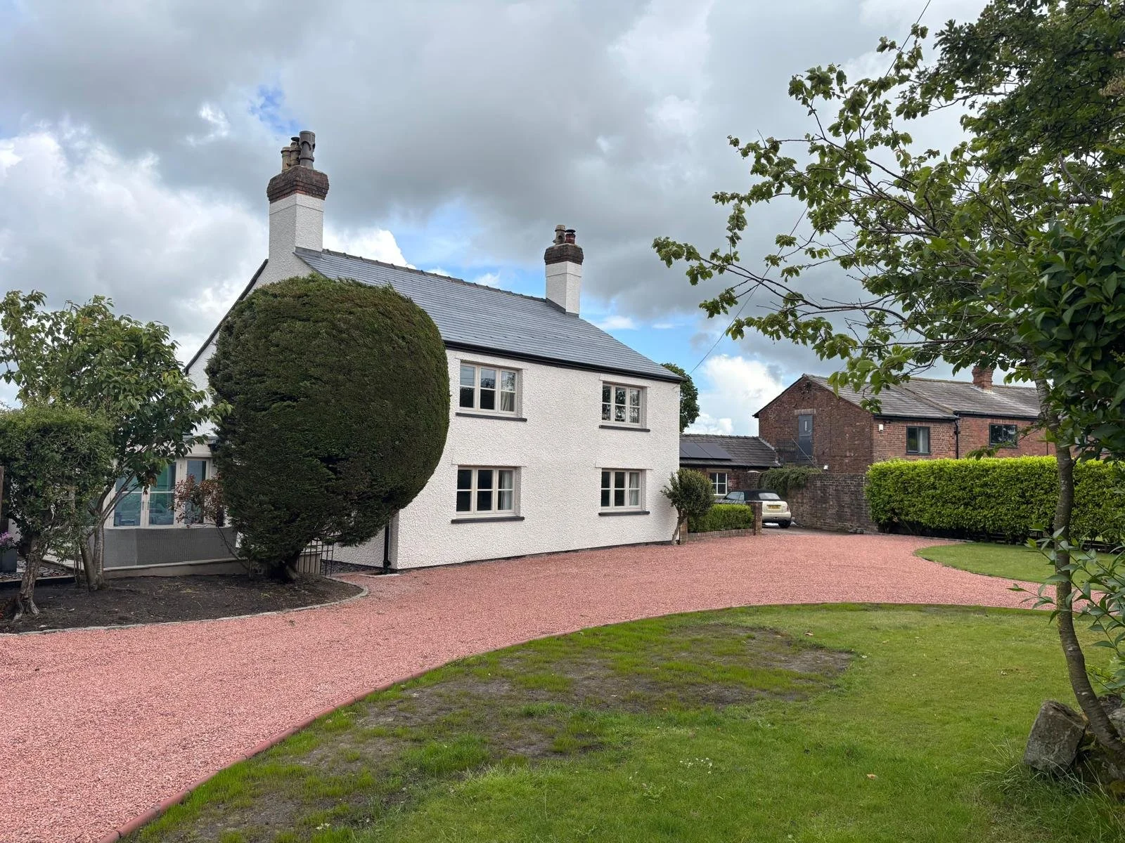 A white house with a metal roof, two chimneys, and several windows, surrounded by a well-maintained garden with trees and shrubs, with a gravel pathway and cloudy sky in the background.