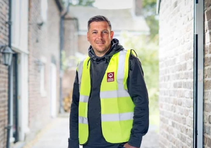 A man wearing a yellow high-visibility vest over a black hoodie, standing in an alleyway with brick walls on either side.