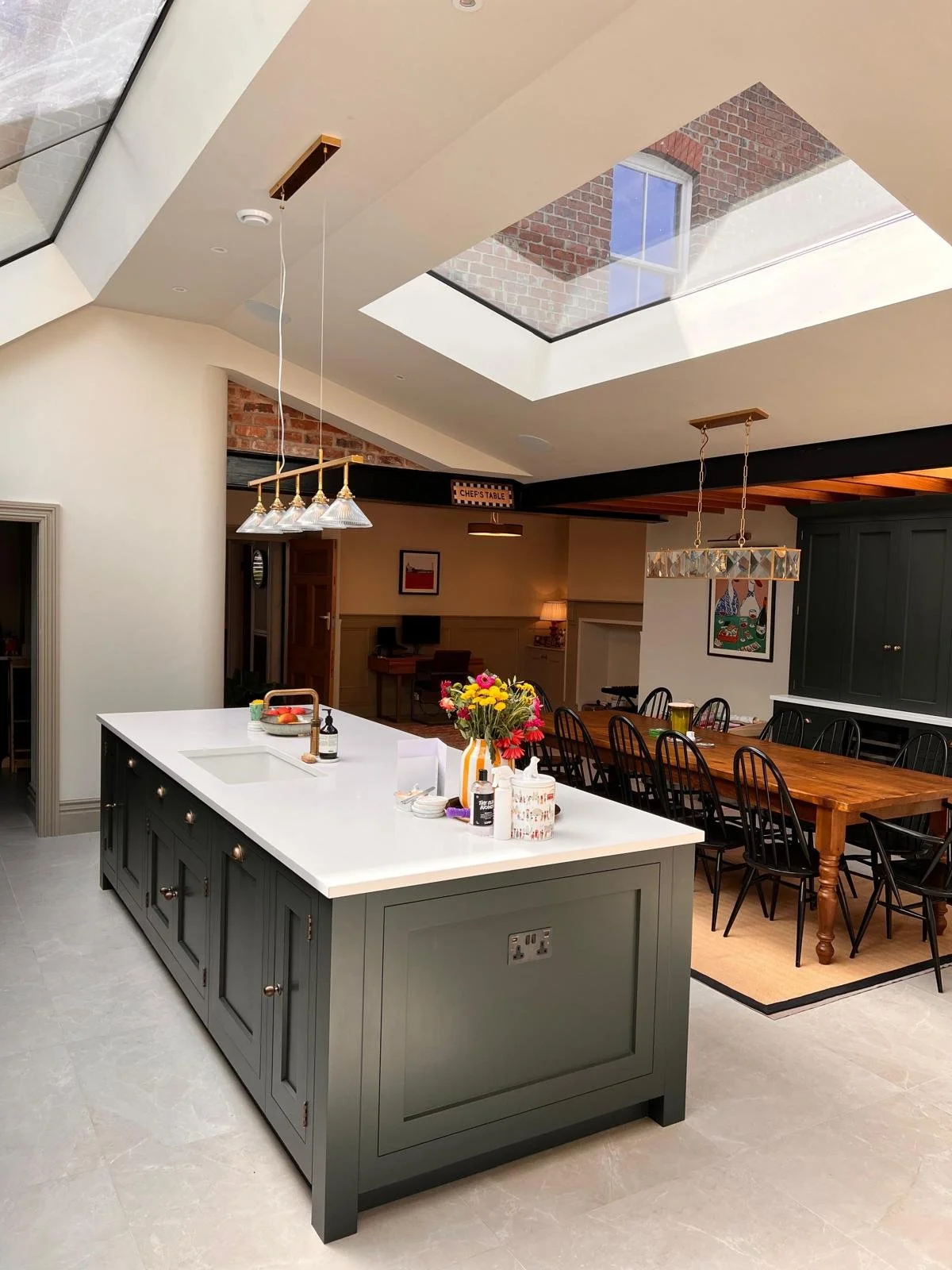 Modern kitchen with a large central island, white countertop, dark green cabinets, and a dining area with a wooden table and black chairs. Skylights, pendant lighting, and exposed brick details are visible.