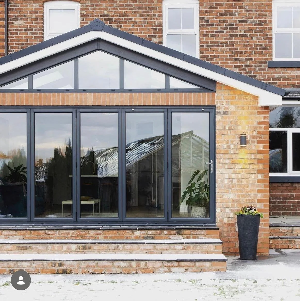 A modern house extension with large glass sliding doors, brick exterior, and a sloped roof, with steps leading up to a patio and a tall black planter with flowers outside.