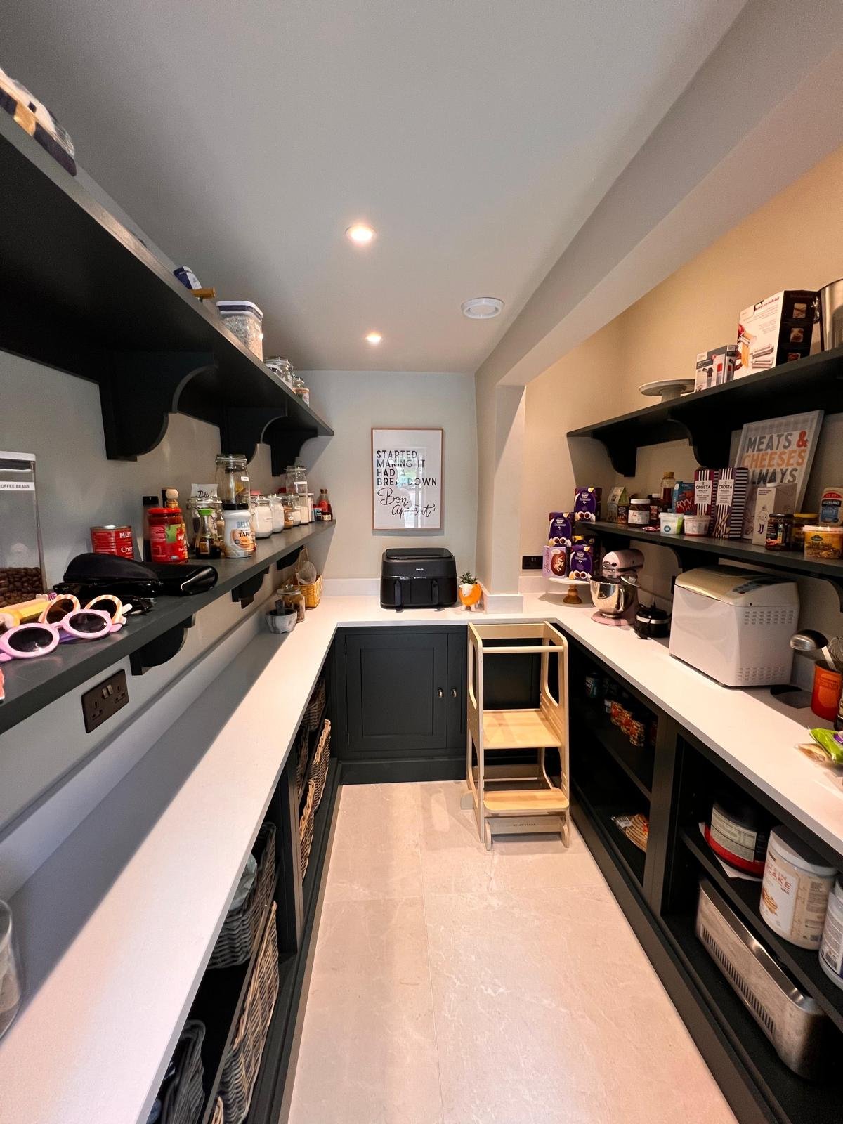 A kitchen pantry with white countertops, black shelves, and various food items, appliances, and kitchen tools organized on the shelves and counters.