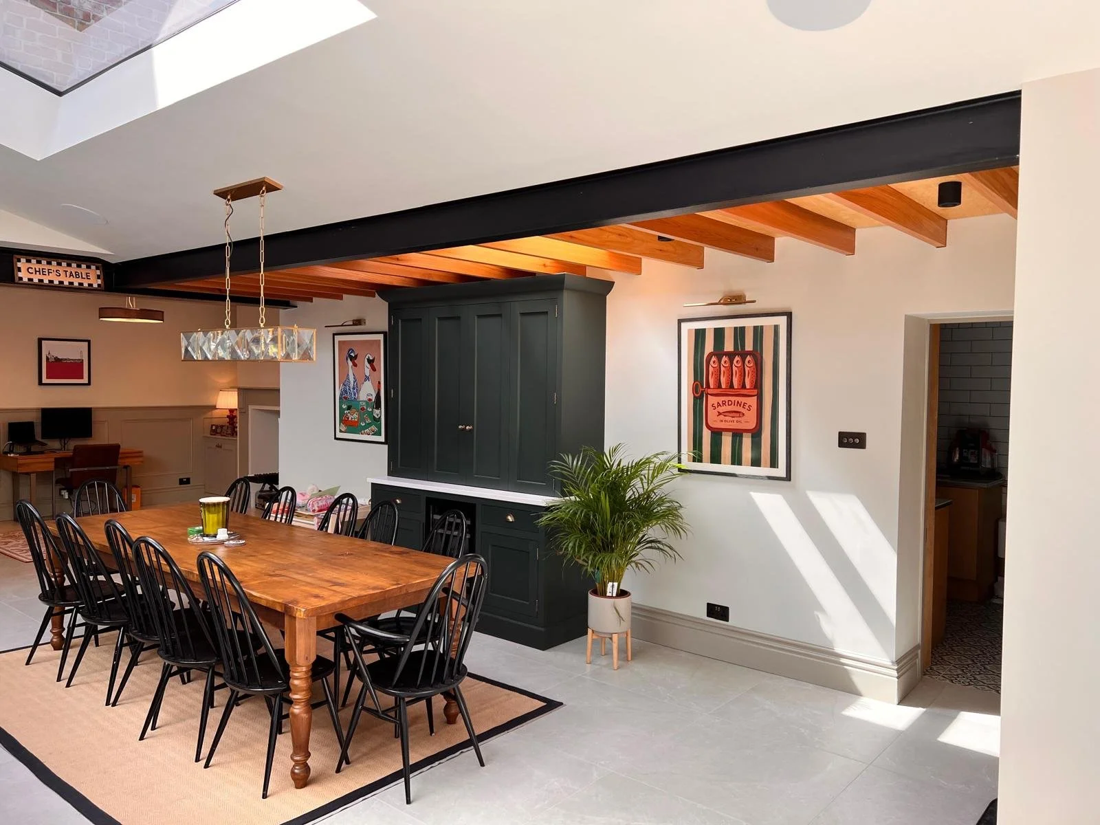 A dining room with a wooden table, black chairs, a green cabinet, wall art, a potted plant, and a skylight letting in natural light.
