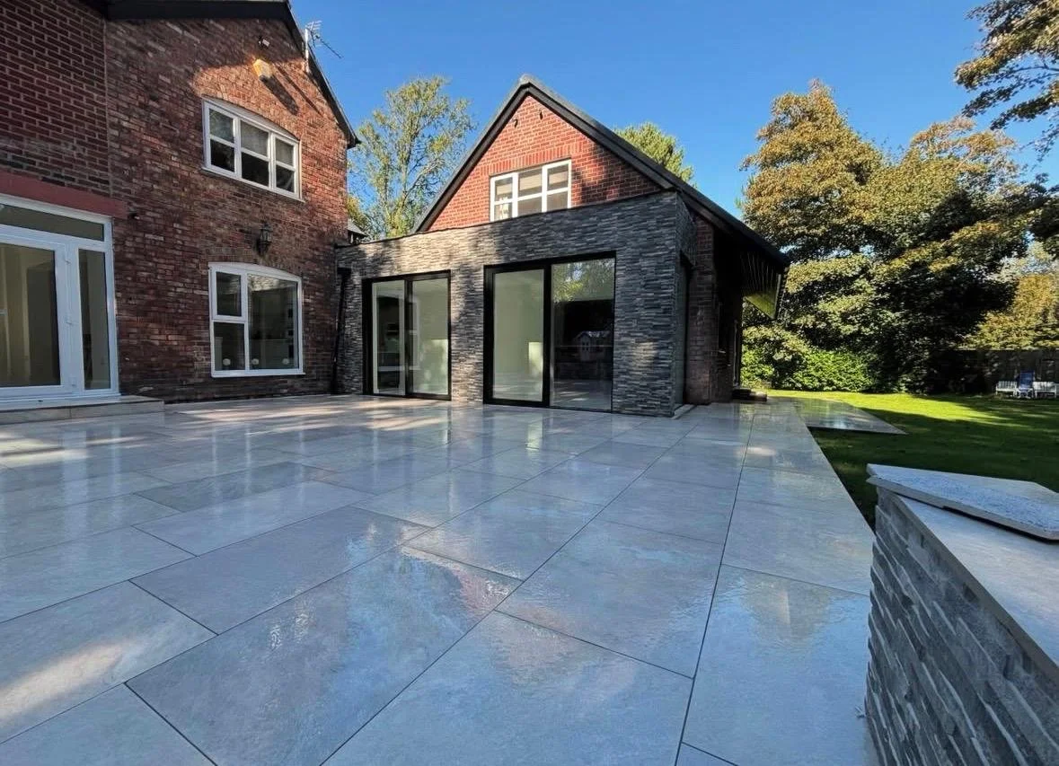 A spacious backyard patio with large white tiles, attached to a brick house with multiple windows and a modern extension with glass doors, surrounded by green trees and a grassy lawn.