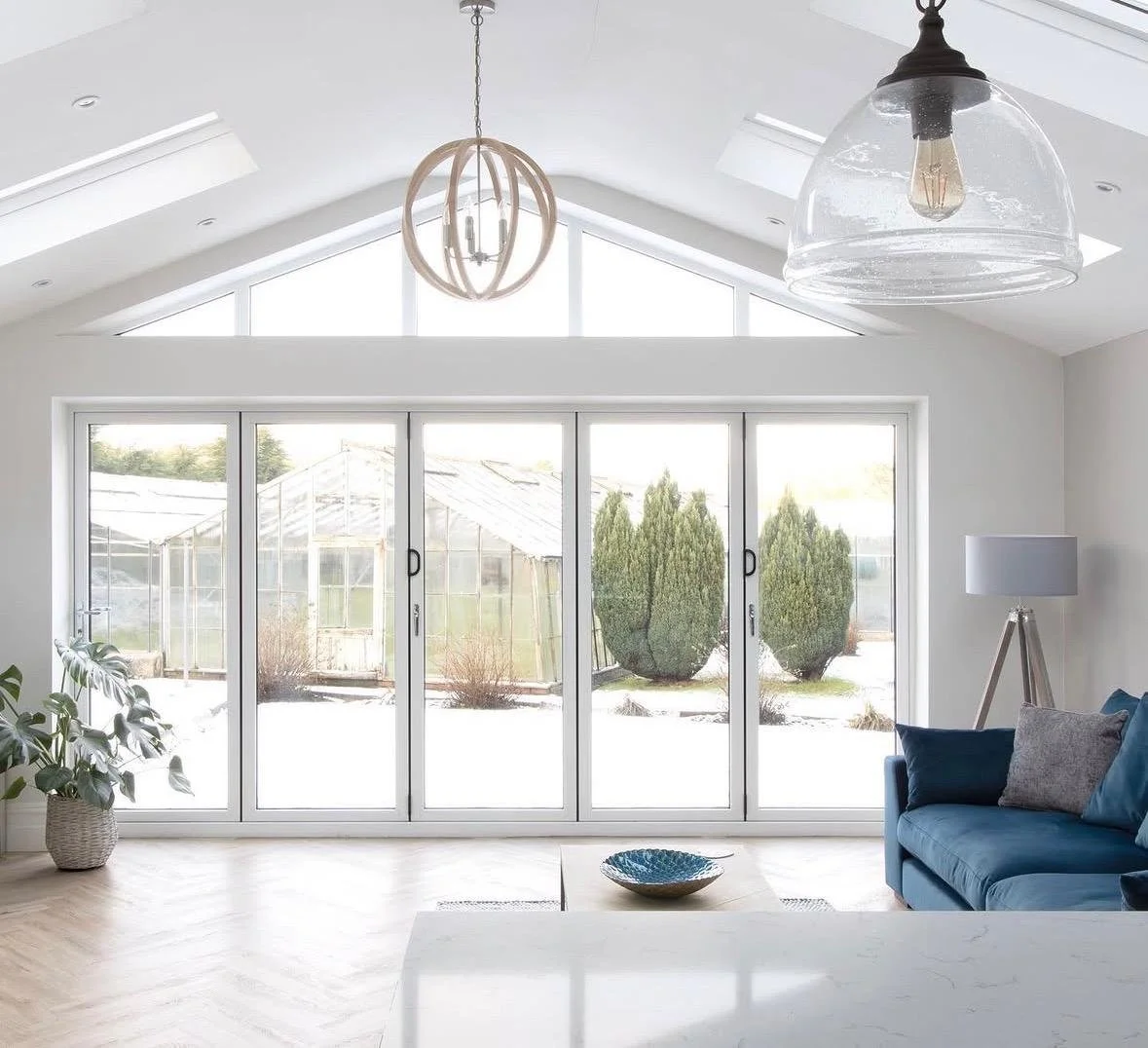 Living room with large sliding glass doors, a blue sofa with pillows, a floor lamp, plants, and hanging light fixtures, overlooking a snowy yard with bushes and a greenhouse.