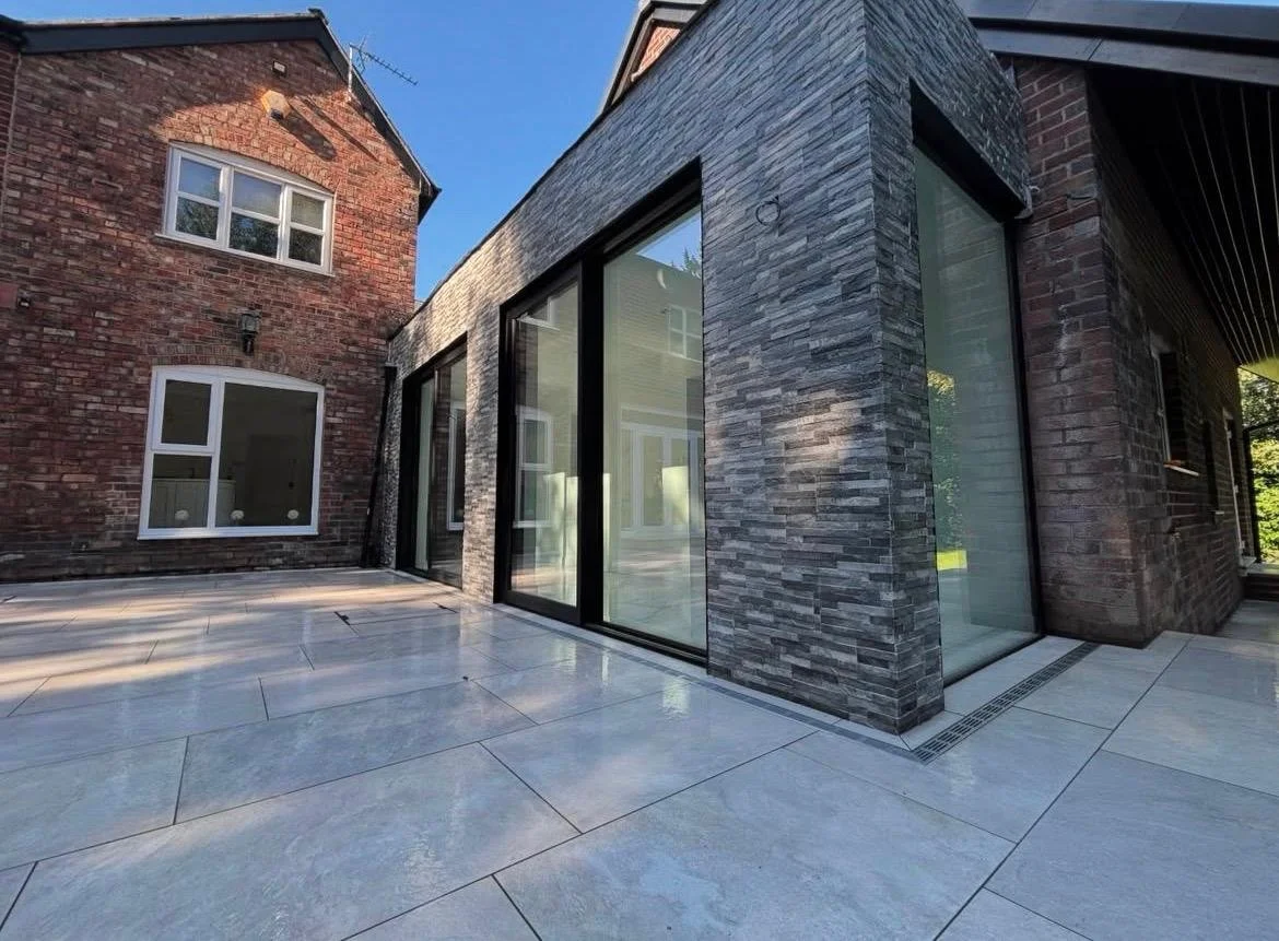 View of a modern building exterior with large sliding glass doors, gray stone and brick walls, and a concrete patio under a clear blue sky.