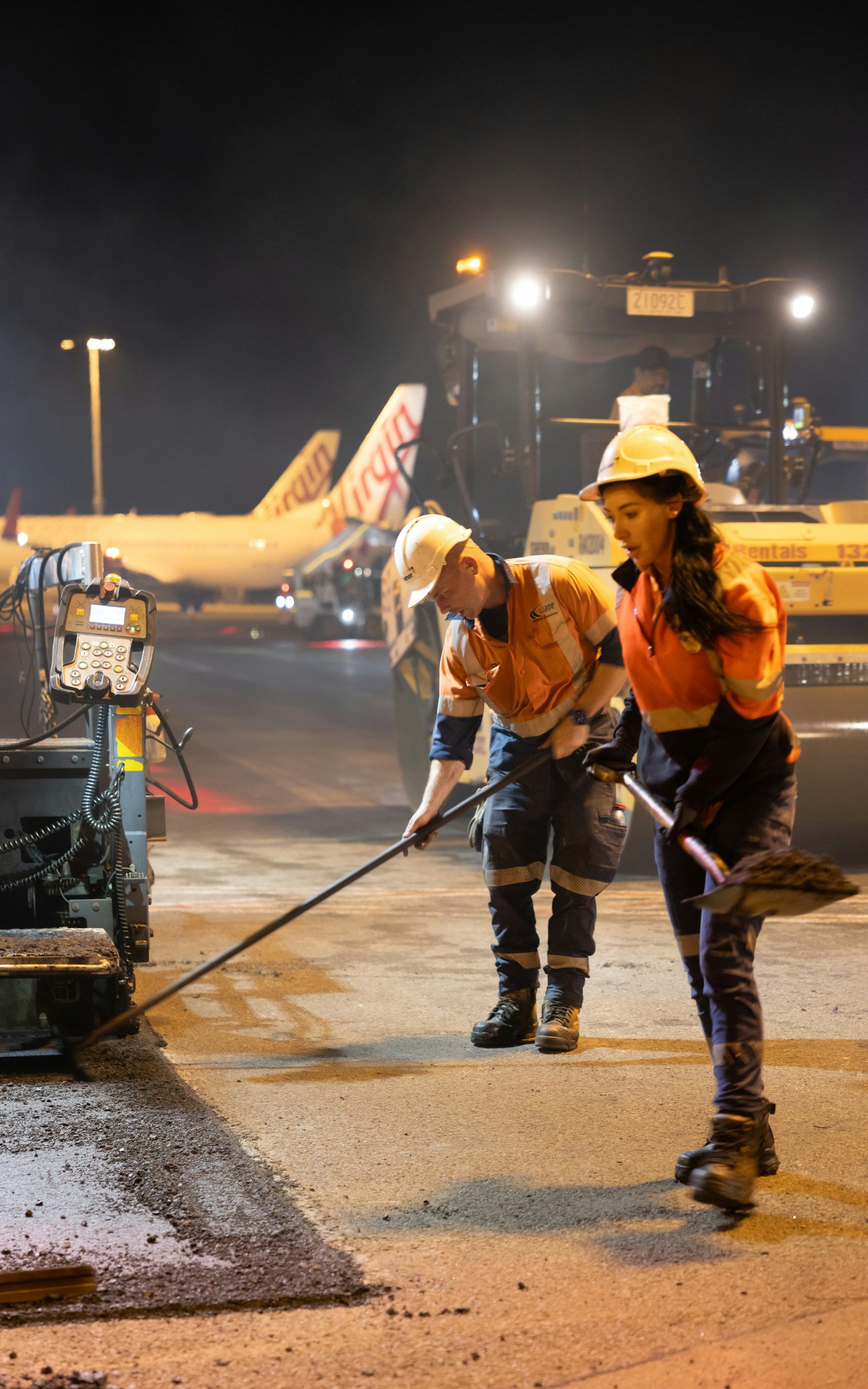 Construction workers wearing safety helmets working on a road at night, with construction equipment and an airplane in the background.