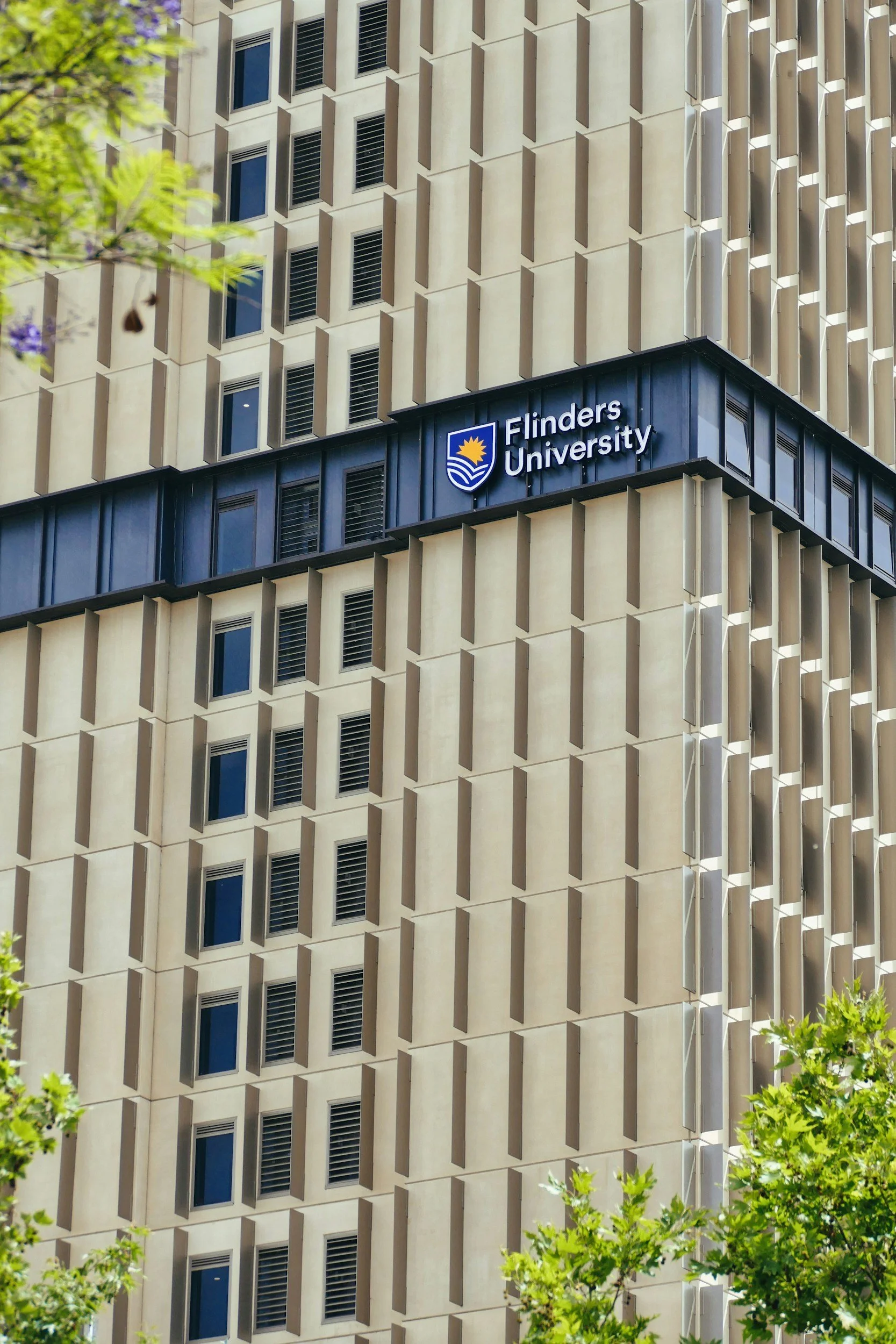 Exterior of a modern university building with windows and vertical sunshade structures, with the sign 'Flinders University' on the corner, surrounded by green trees.