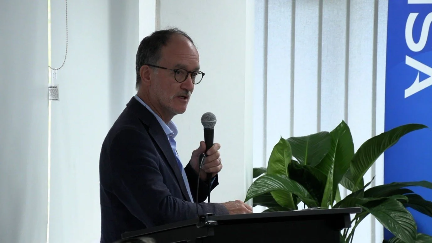 A man wearing glasses and a dark blazer standing at a podium holding a microphone, with a large green plant nearby and part of a blue sign with white text on a white wall in the background.