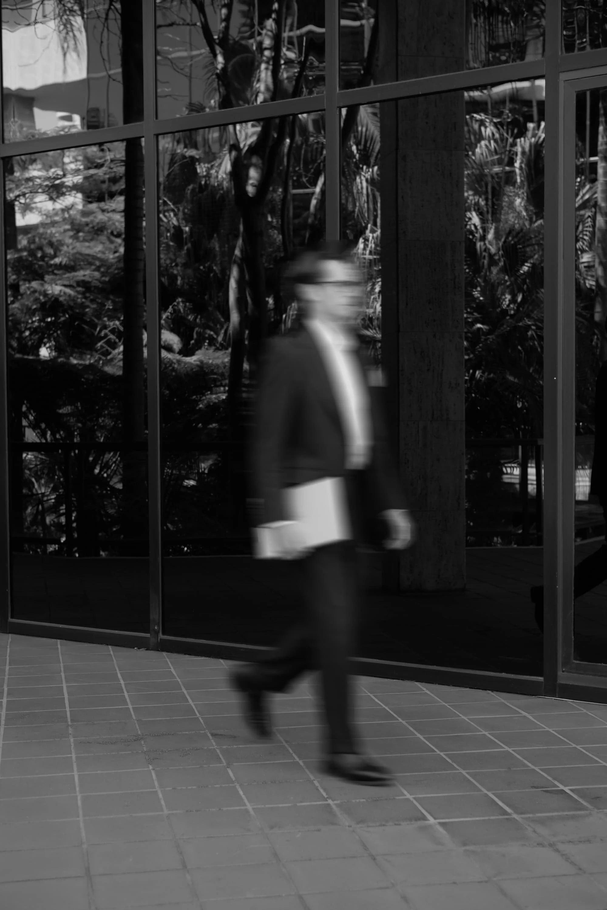 A blurred person walking inside a modern building with large glass windows and potted plants outside.