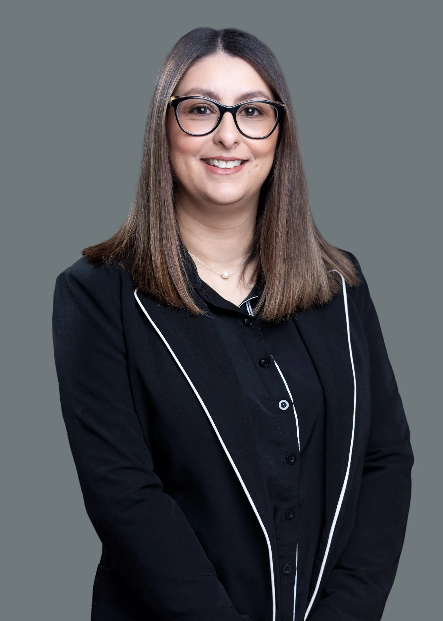 A woman with straight, shoulder-length brown hair, wearing glasses and a black blazer with white trim, smiling in front of a plain gray background.