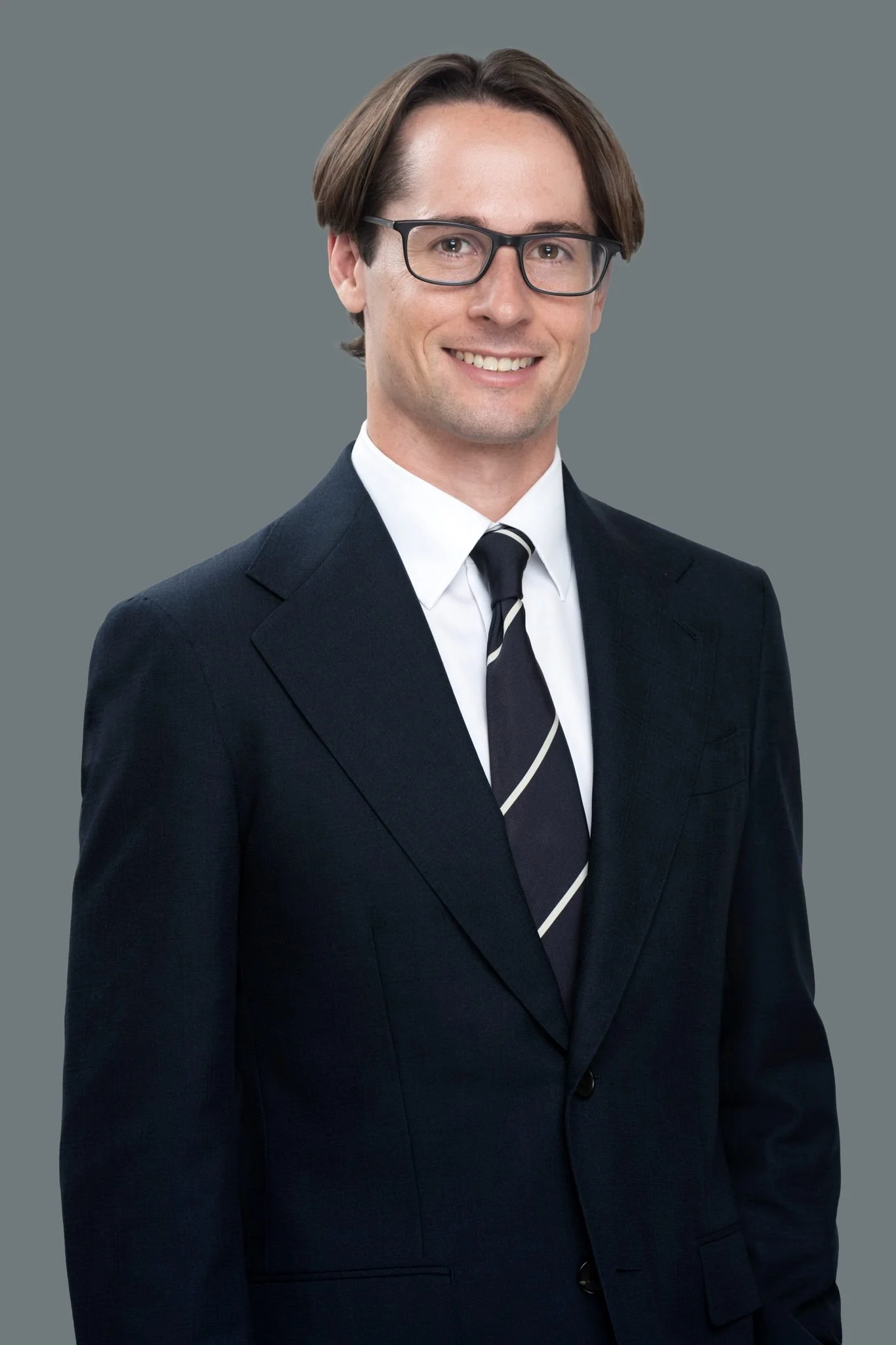 A man in a business suit, white shirt, and striped tie, smiling, wearing glasses, with a neutral gray background.