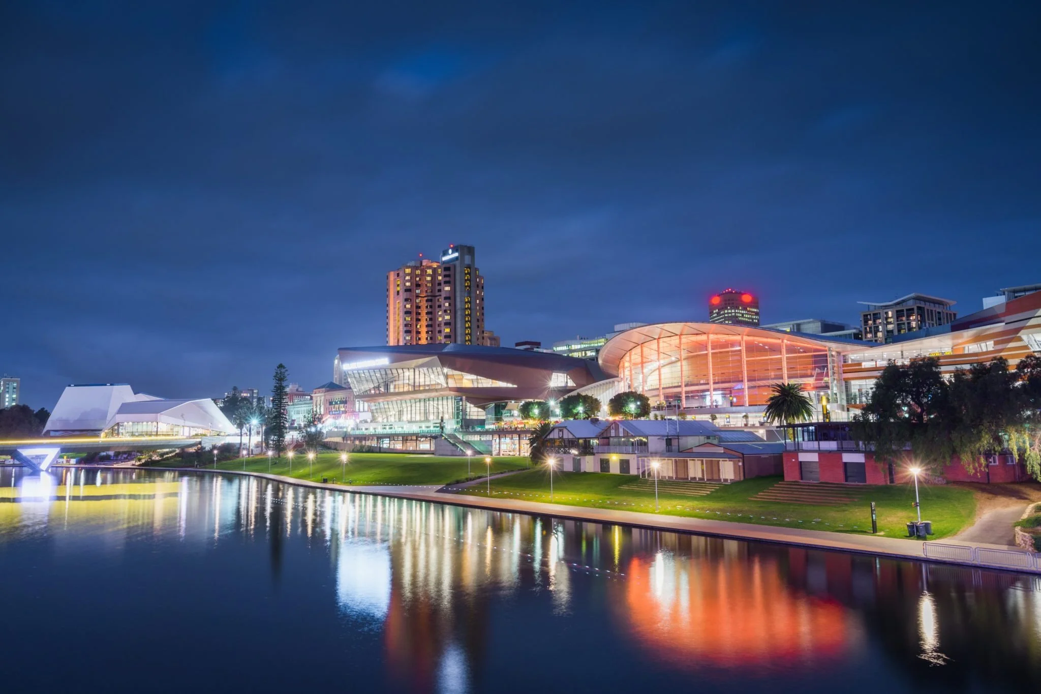 Night view of a city skyline with illuminated buildings, a waterfront with reflections, and a modern architectural structure, under a dark cloudy sky.