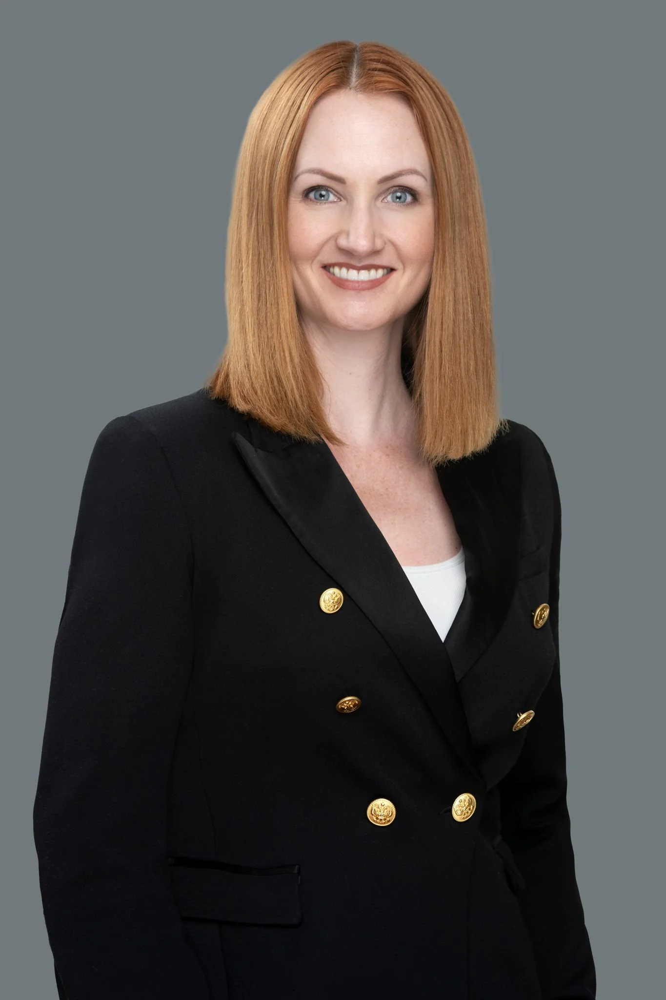 A woman with straight red hair, blue eyes, and fair skin, wearing a black blazer with gold buttons against a gray background.