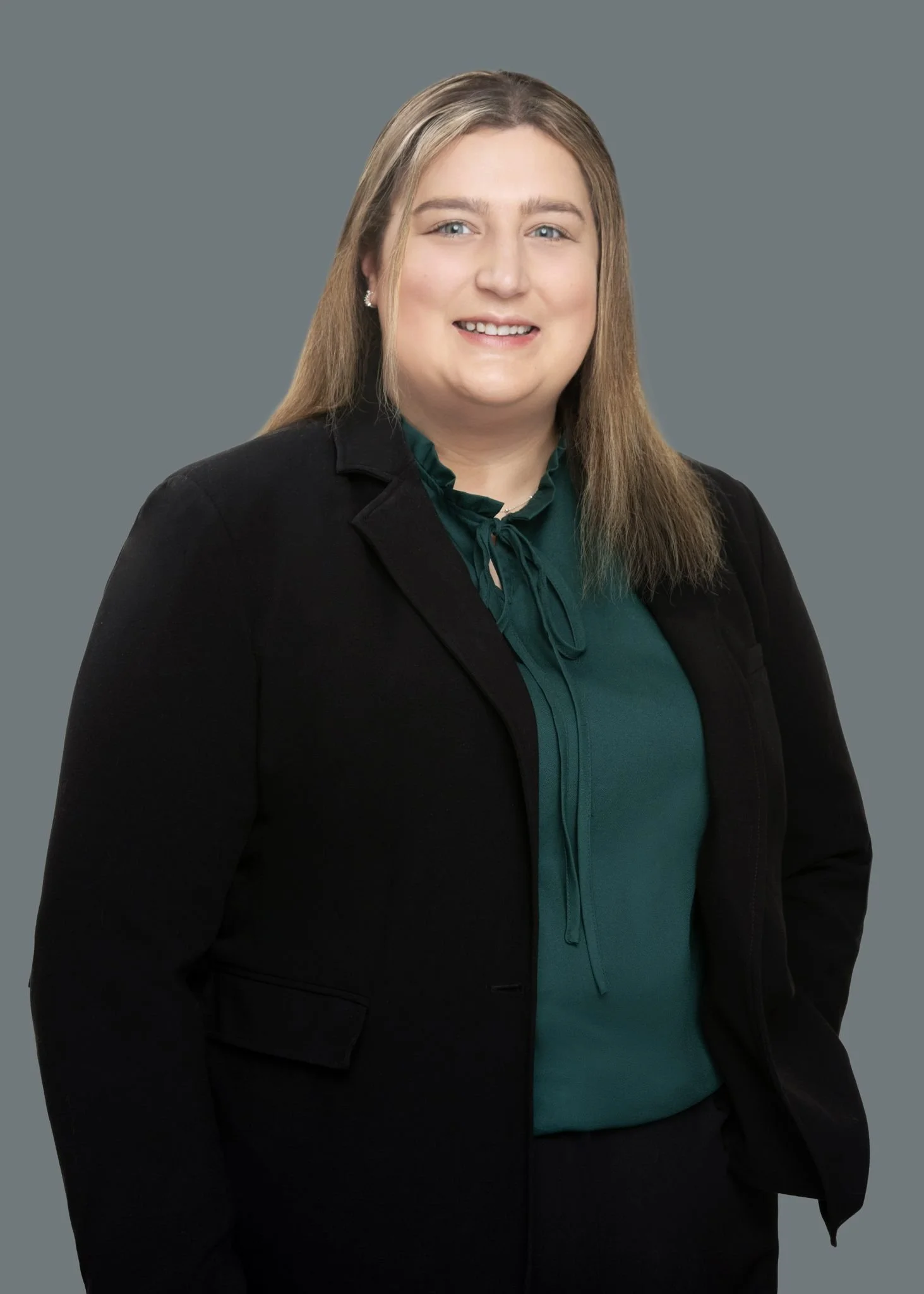 A professional woman with light brown hair, wearing a dark blazer and a teal blouse, standing against a gray background.