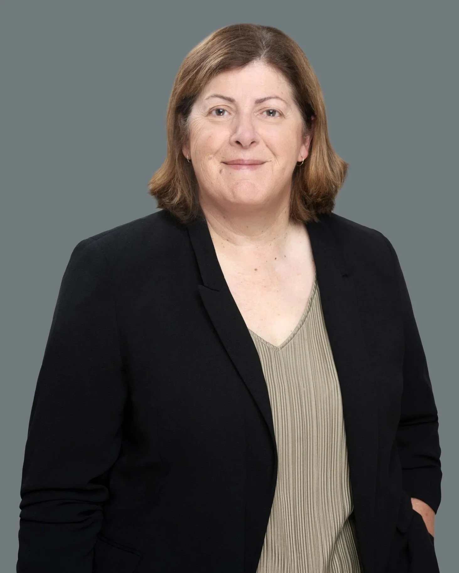 A professional headshot of a woman with shoulder-length light brown hair, wearing a black blazer over a beige ribbed top, smiling slightly against a plain gray background.