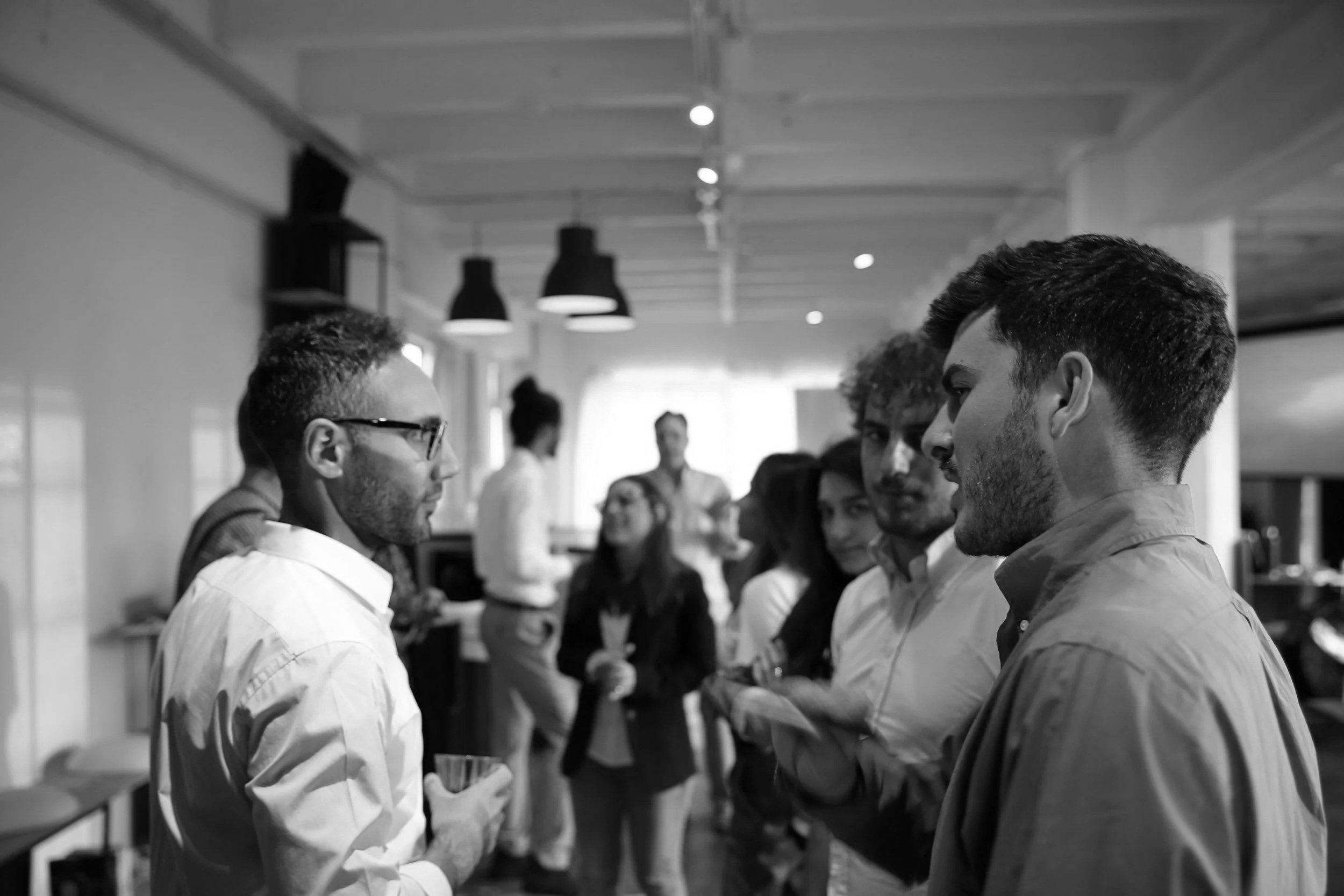 Black and white photo of two men with short hair and glasses or no glasses each, standing face to face and engaging in conversation in a modern cafe or restaurant, with other people in the background. The lighting includes modern ceiling lamps.