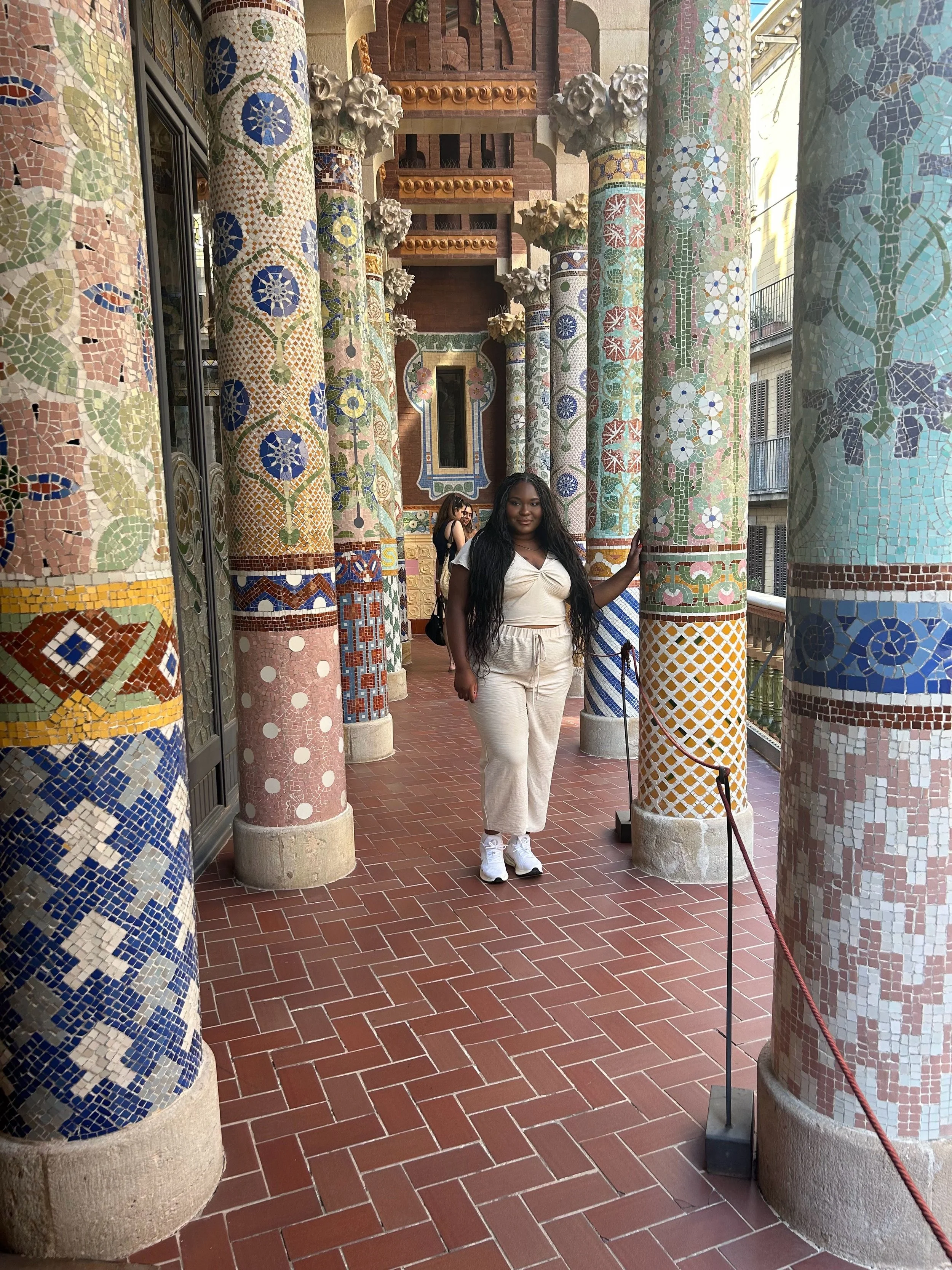 Young woman with long black hair, wearing a white top and white pants, stands among decorative mosaic-covered columns in a historic setting, with other visitors in the background.