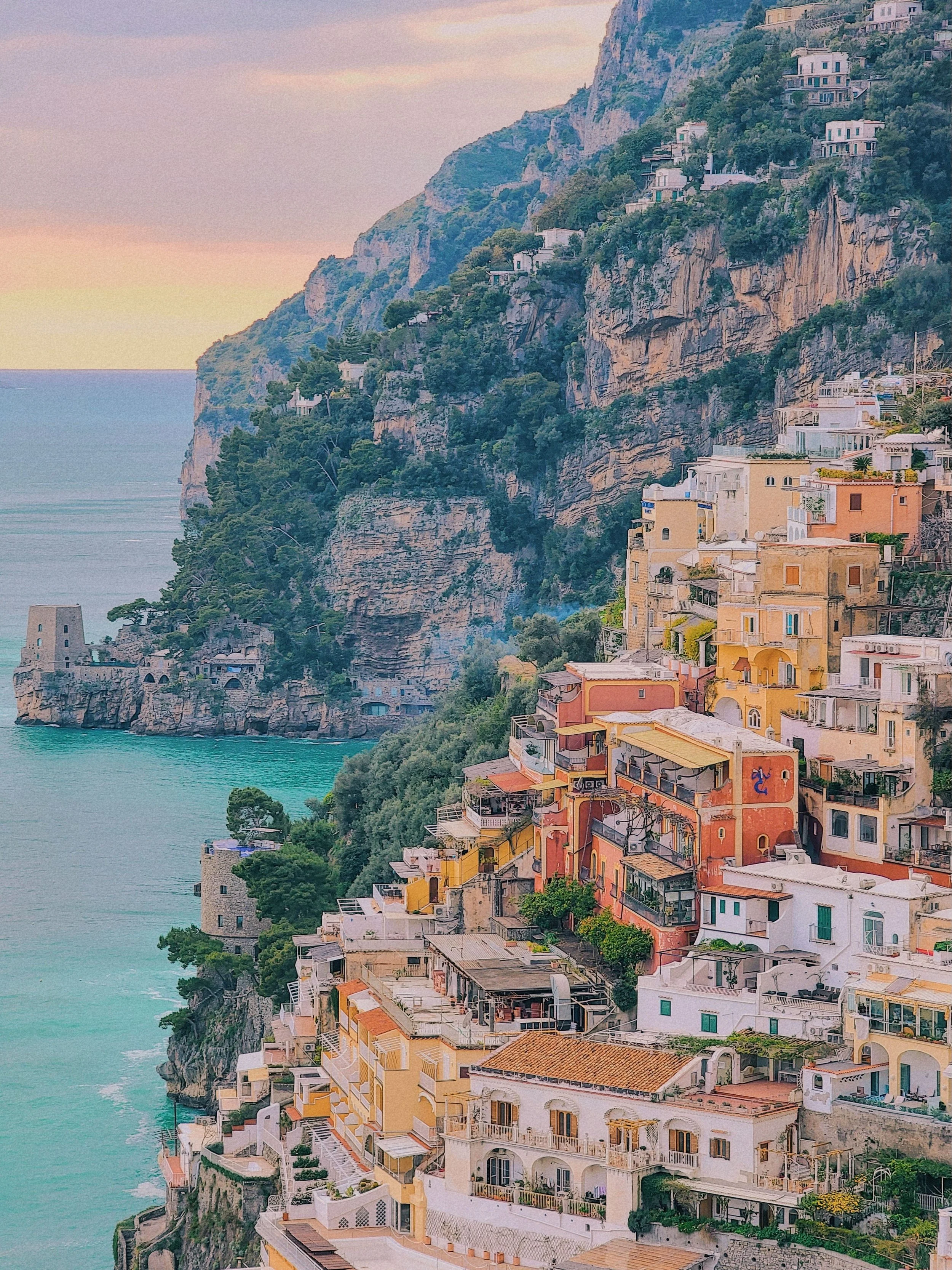 Colorful seaside town built into cliffs, with pastel-colored buildings and lush greenery overlooking the ocean at sunset.