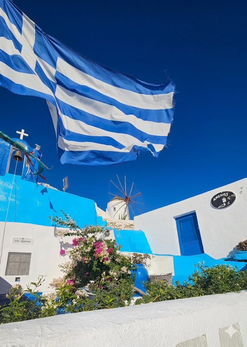 Blue and white Greek flag waving against a clear blue sky, with white buildings, a windmill, and colorful flowers in the background.