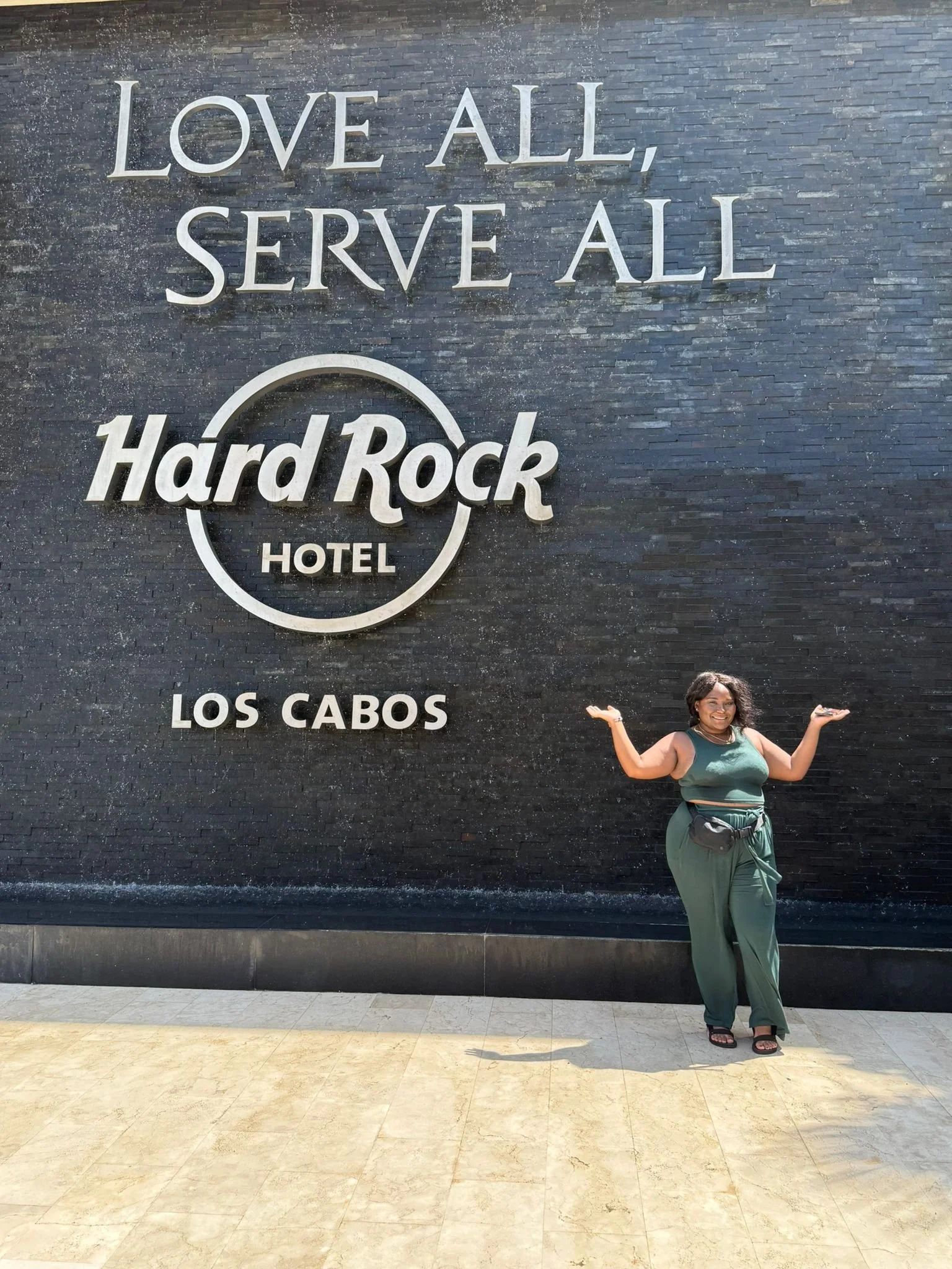Woman standing in front of a wall with the sign 'LOVE ALL, SERVE ALL, Hard Rock Hotel, LOS CABOS', gesturing with both hands raised near her shoulders.