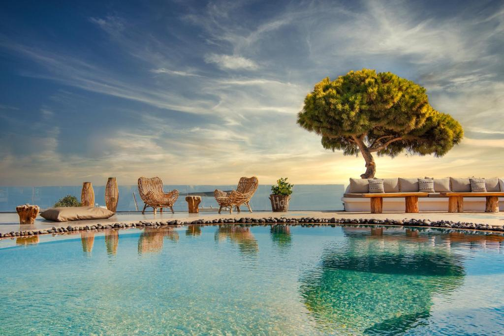 A serene seaside outdoor pool area with wicker chairs, a large tree, and cushioned benches, overlooking the ocean at sunset.
