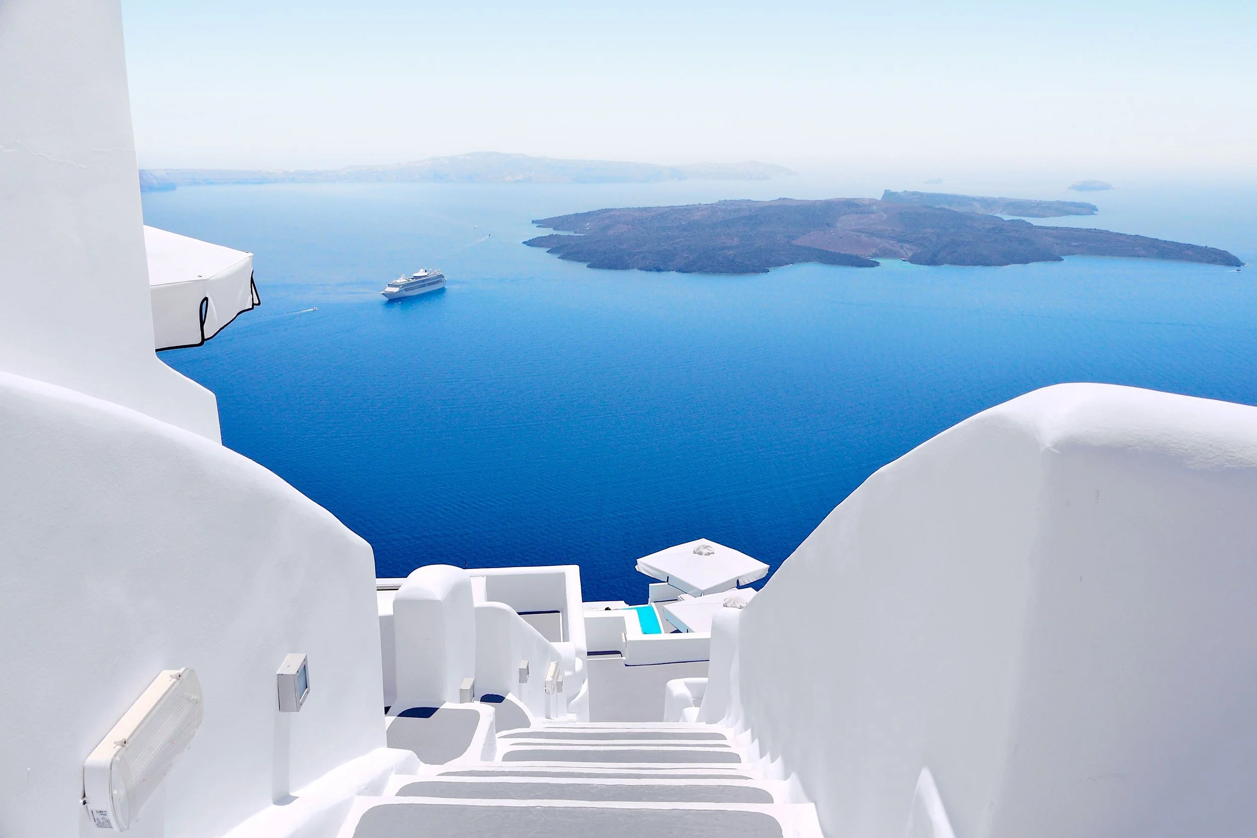 White buildings with curved surfaces and stairs overlooking deep blue sea, cruise ship sailing near islands in the distance, clear sky, typical of Santorini Greece.