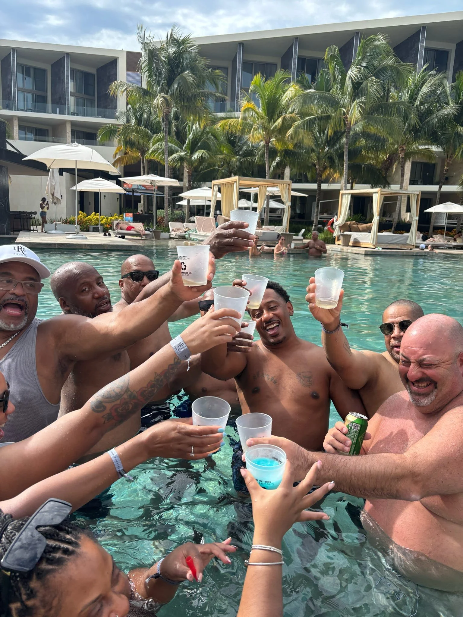 Group of people enjoying a pool party, raising drinks in celebration, with a resort or hotel in the background, featuring palm trees and lounge beds.