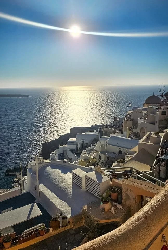 Sun setting over the ocean, casting a shimmering reflection on the water, with white buildings and terraces on a hillside in a Mediterranean coastal town.