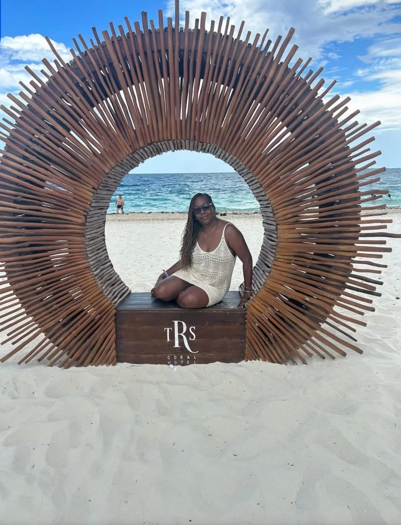 A woman sitting on a wooden platform inside a large circular bamboo structure on a sandy beach, with the ocean and a partly cloudy sky in the background.