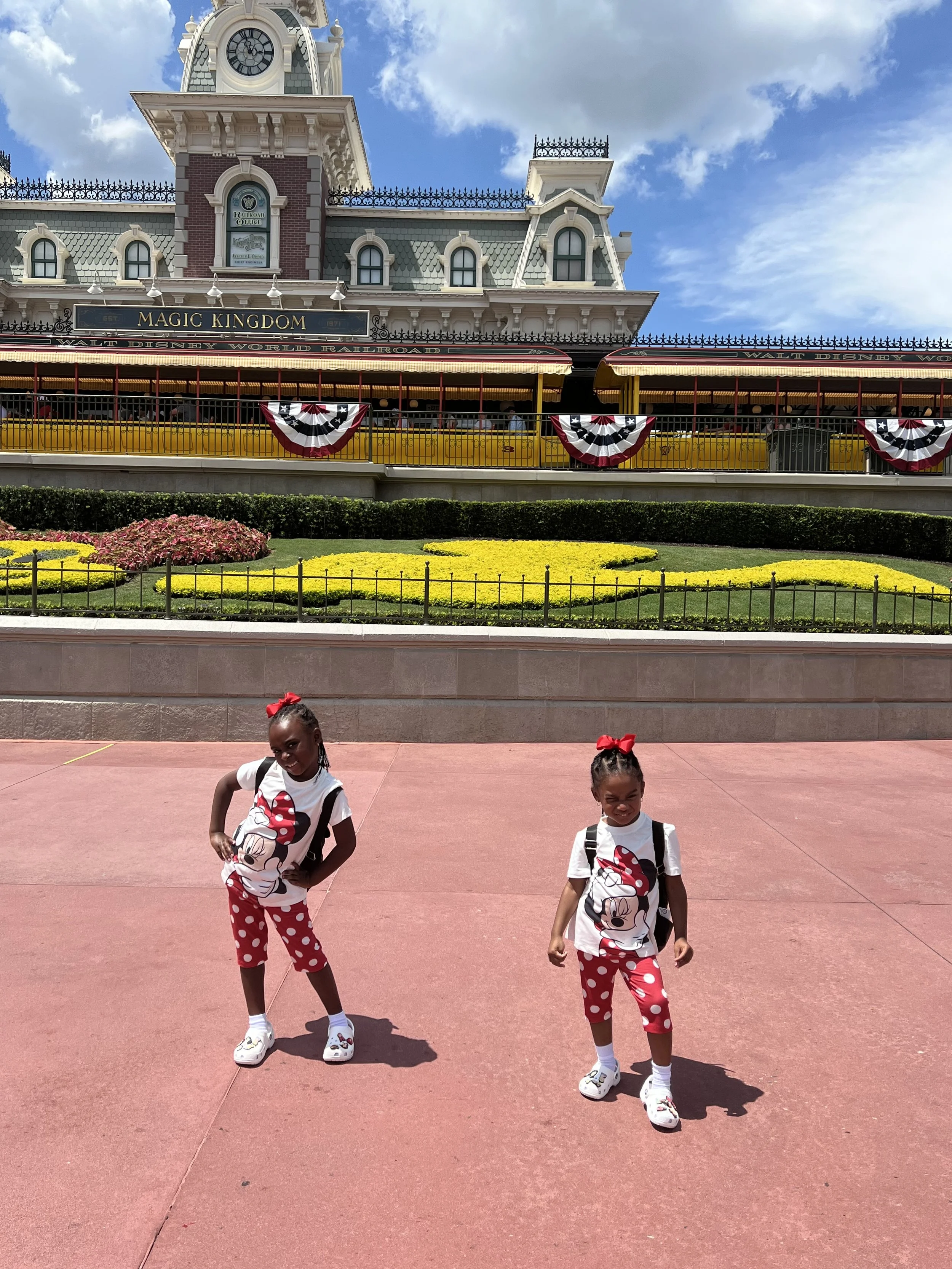 Two young girls standing on a pinkish-red pavement in front of Disneyland's Main Street, USA entrance decorated with red, white, and blue bunting, with a large building resembling a train station and a yellow train in the background.