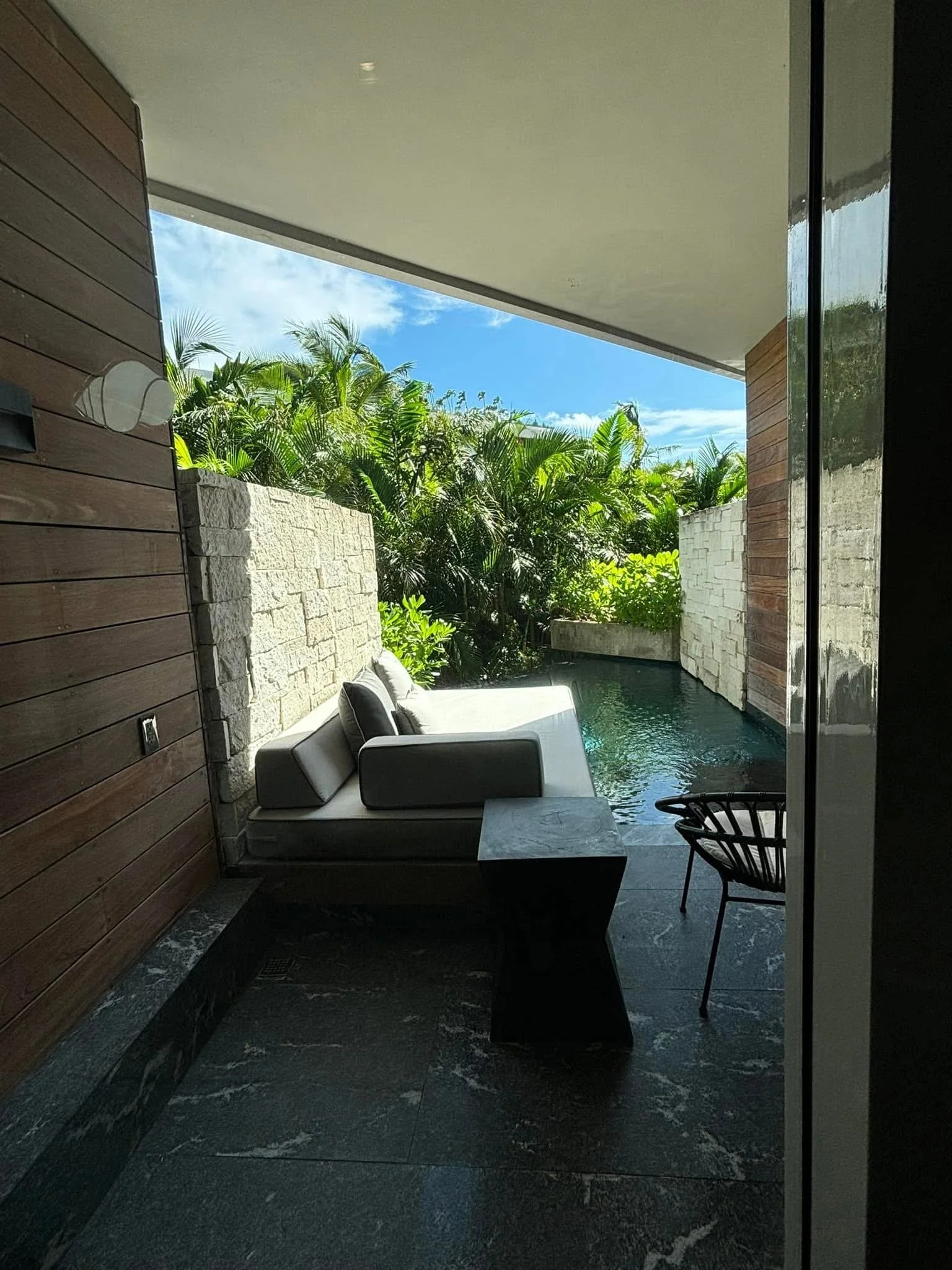 Indoor lounge area with a black stone floor, a white sofa with cushions, a dark small side table, a chair, a wooden wall, and a large window showing lush green tropical plants and a blue sky.