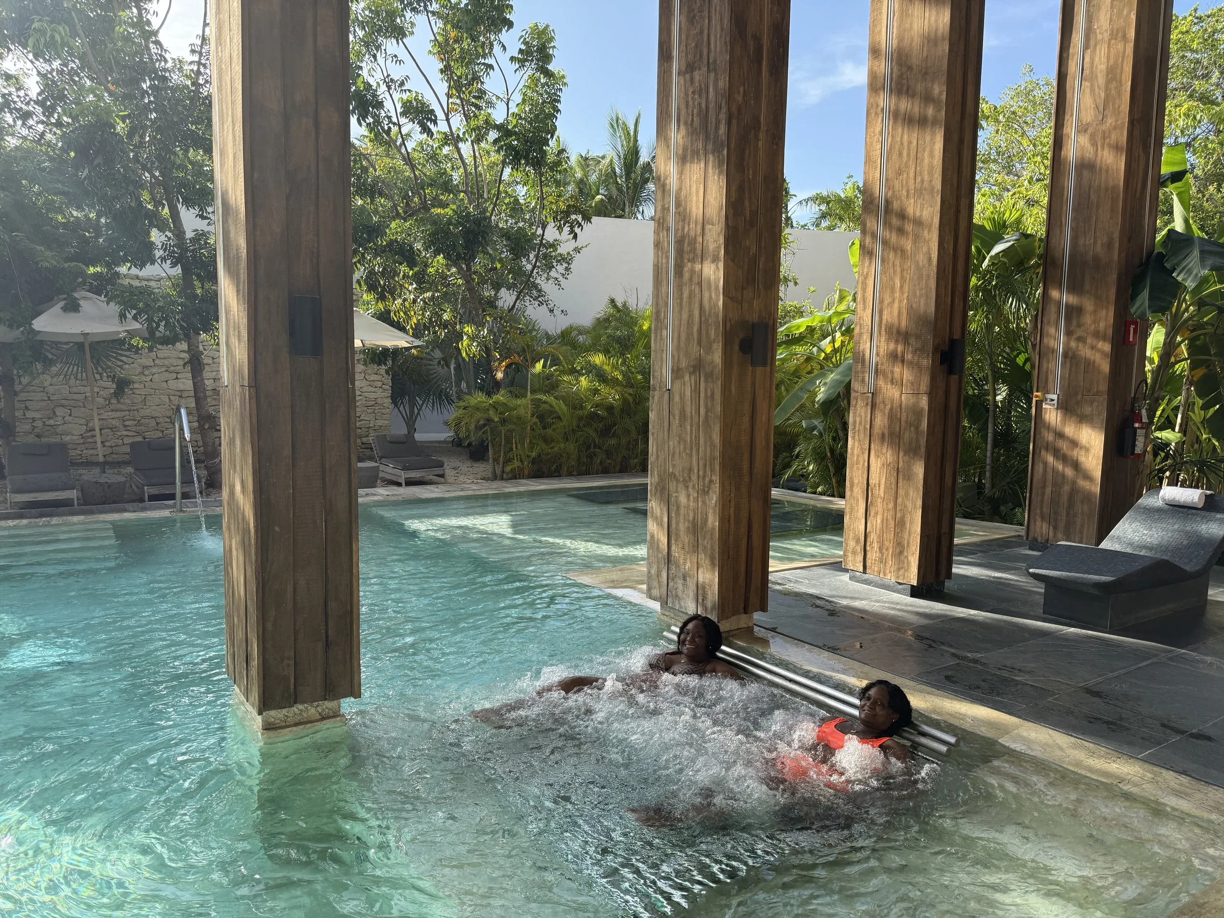 Two women enjoy a hot tub or pool outside with lush greenery and trees surrounding the area, under a sunny sky.