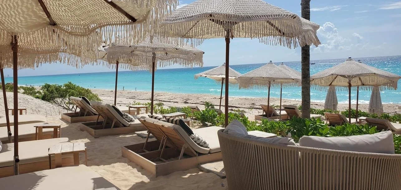 Beach scene with white sand, turquoise water, palm trees, white umbrellas, and wooden lounge chairs under umbrellas