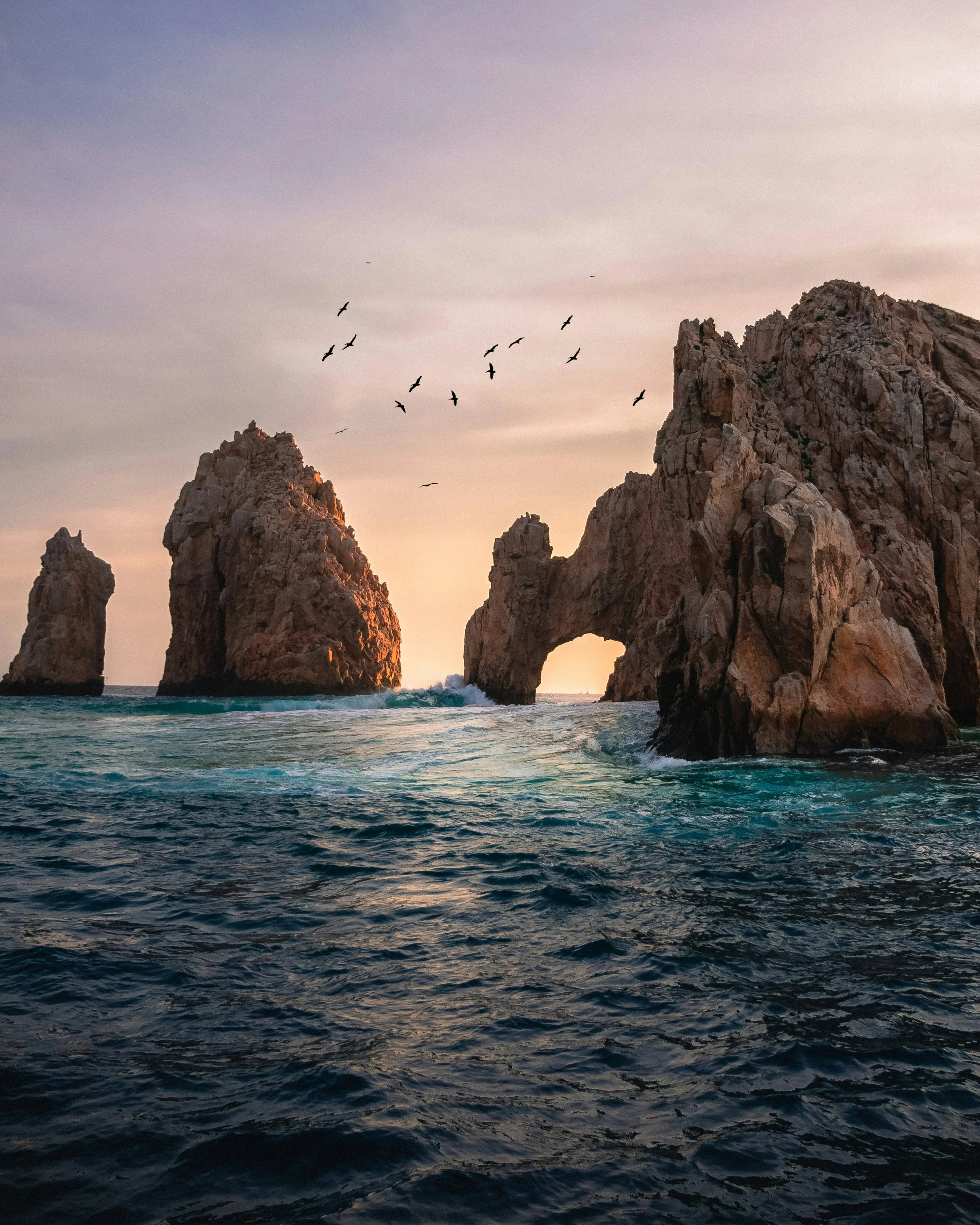 Seascape with rocky formations rising from the ocean and a natural rock arch in the background during sunset, with a flock of birds flying in the sky.
