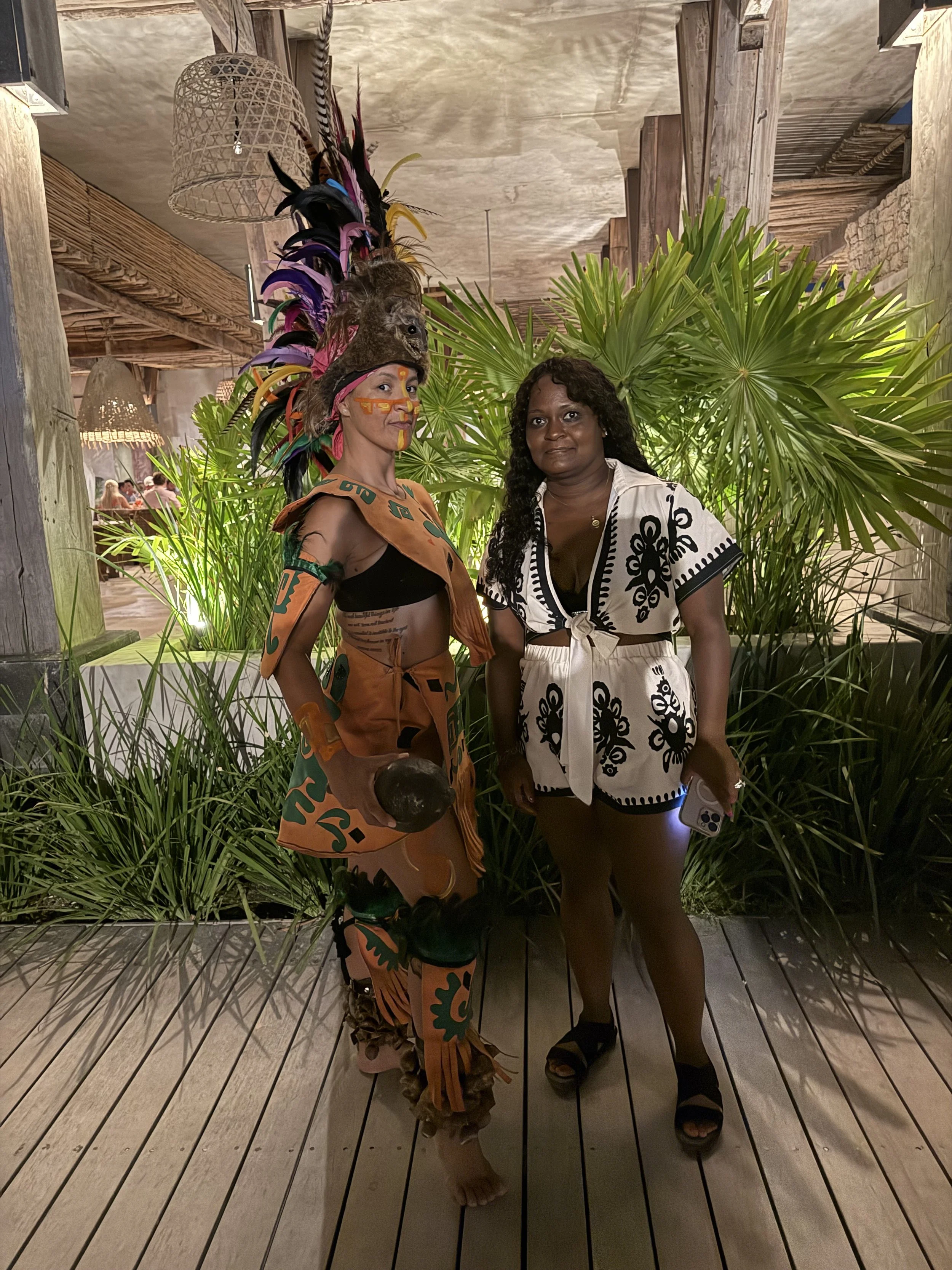 Two women standing on a wooden deck with greenery in the background, one dressed in traditional tribal attire with a feathered headdress, face paint, and earthy accessories, and the other in a black and white printed summer outfit holding a smartphon