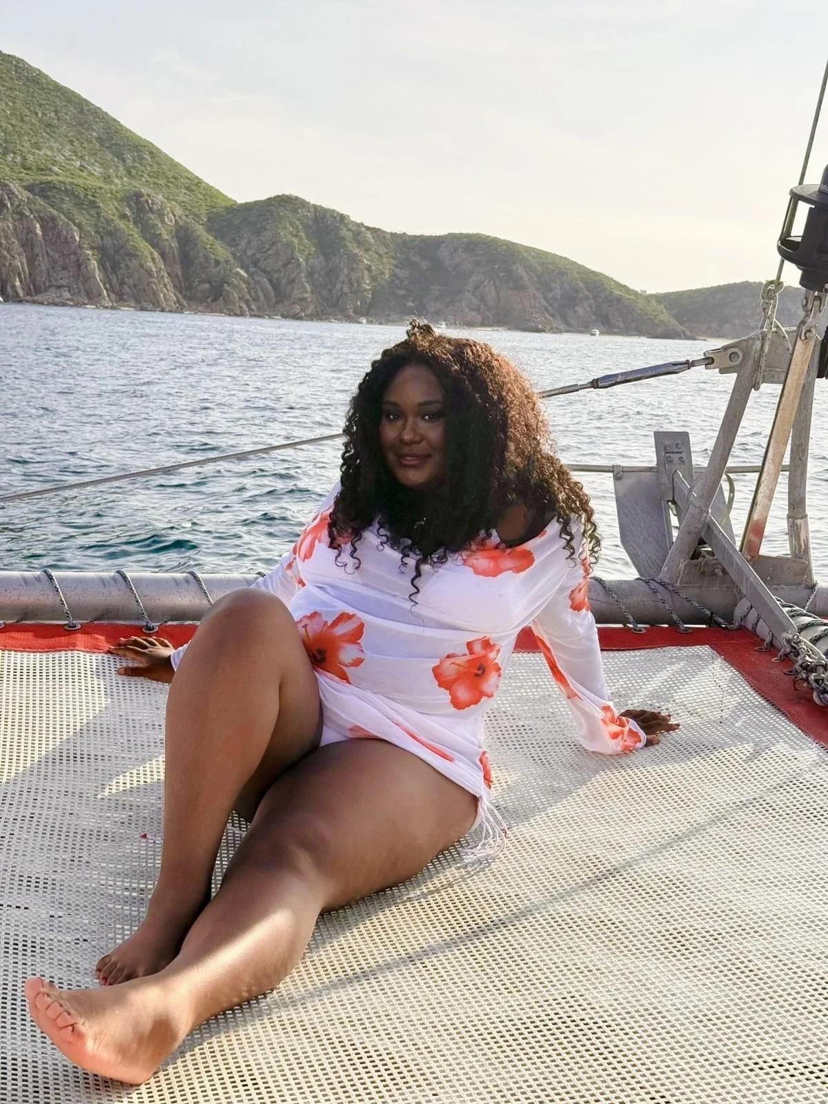 A woman with curly hair sitting on a boat deck near the water, wearing a white dress with red floral patterns, with a landscape of hills and water in the background.