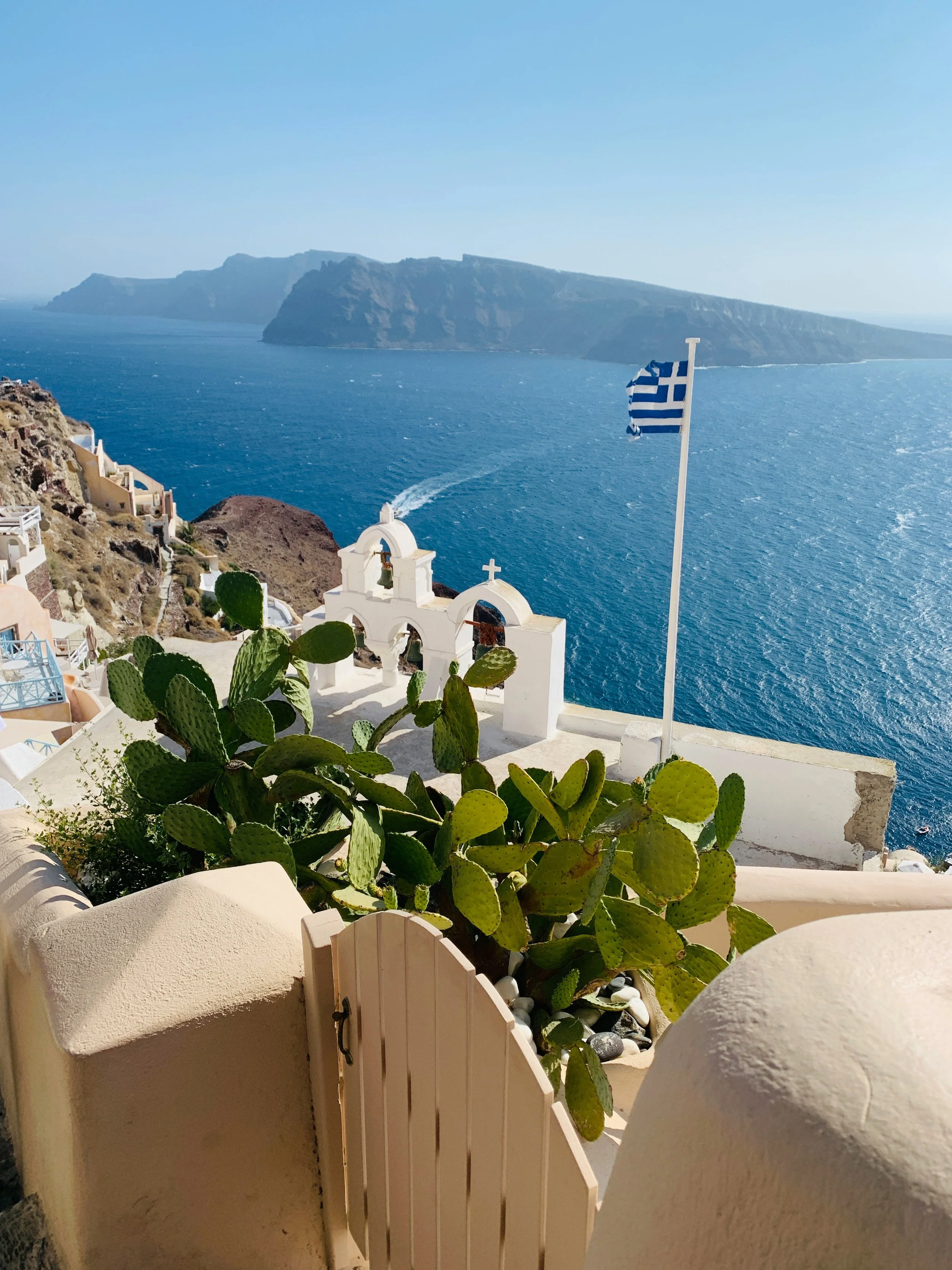 A scenic view of a coastal village with white buildings, a flagpole with a Greek flag, and the ocean with cliffs in the background, suggesting a Mediterranean setting.